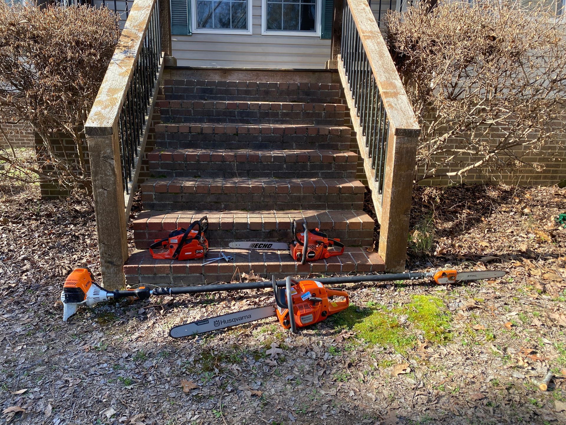Chainsaws and other yard tools sit on a porch and ground, ready for use. The tools are orange and silver.