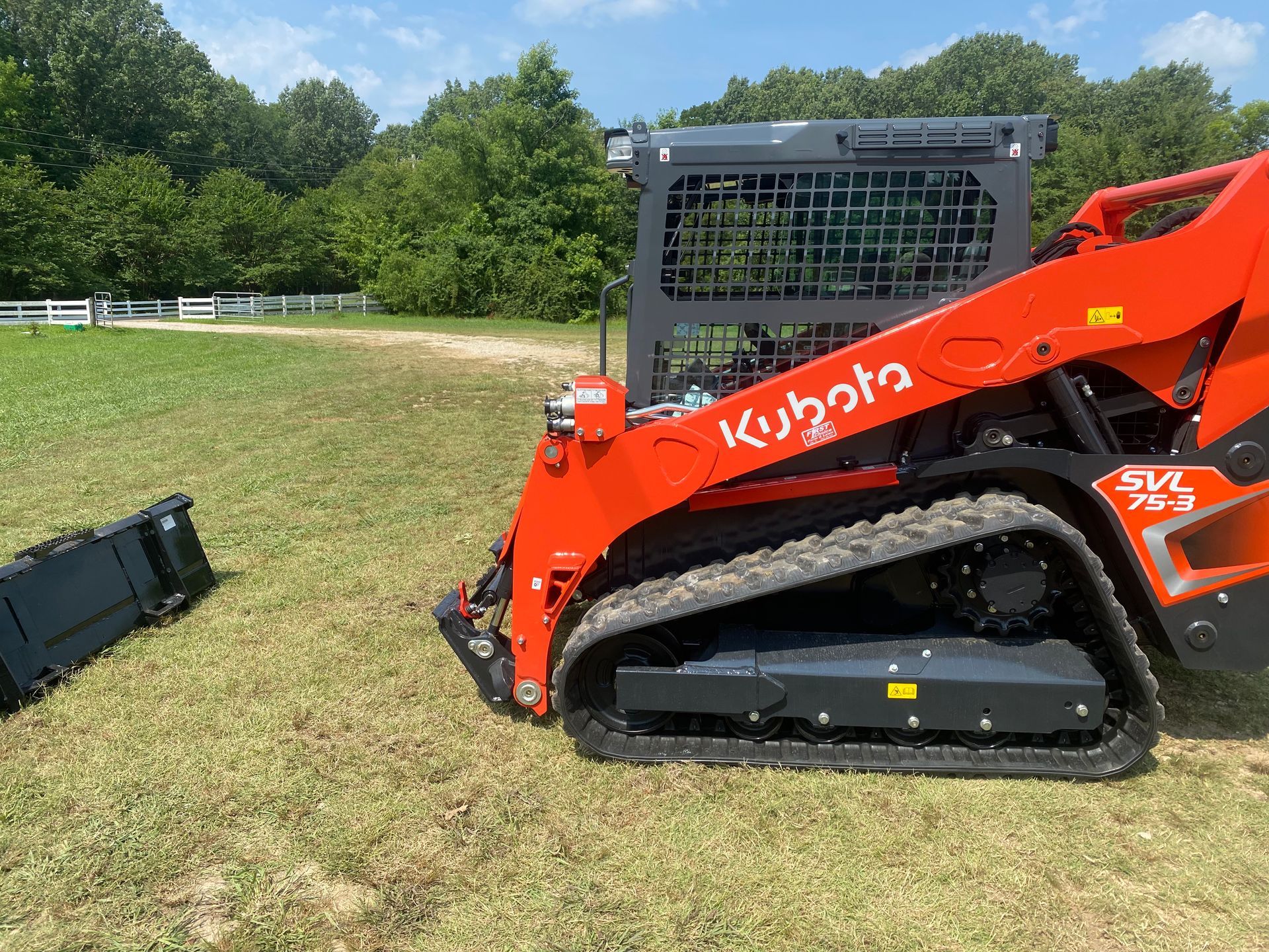 A bright orange Kubota skid steer loader with tracks, next to a detached black bucket, on a grassy field.