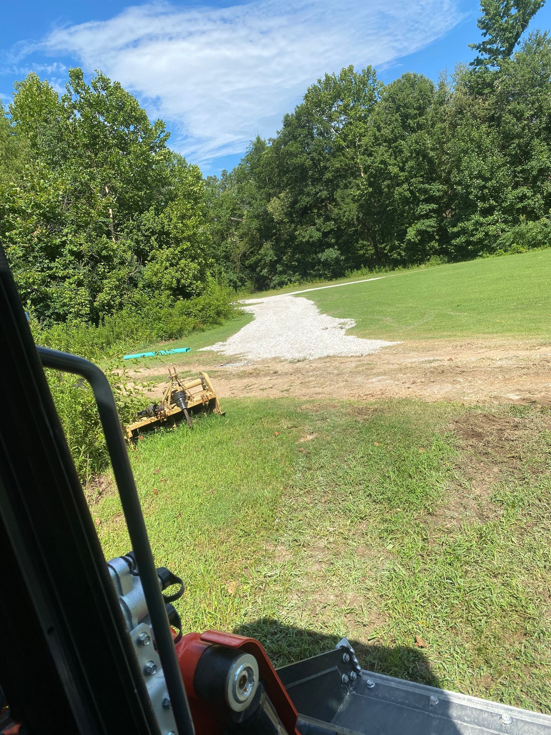 A pile of gravel on a grassy clearing surrounded by trees, viewed from inside a small construction vehicle.