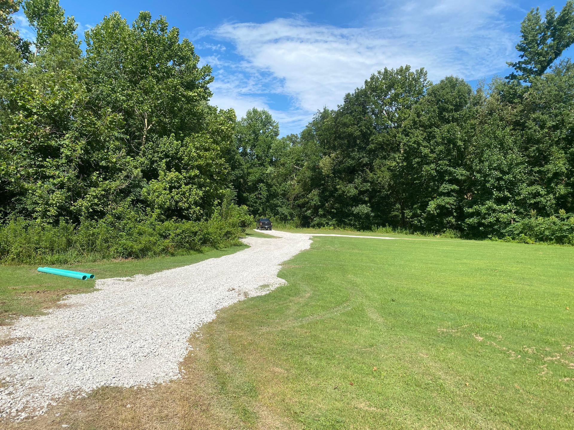 Gravel driveway leading into trees, with a green lawn on the right. Blue sky with clouds overhead.