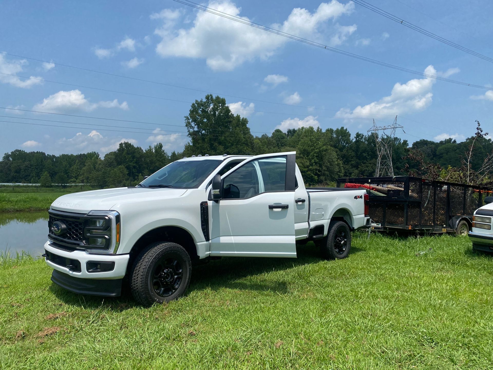 White pickup truck with a black grill and wheels parked on green grass near water, with the door open on a sunny day.