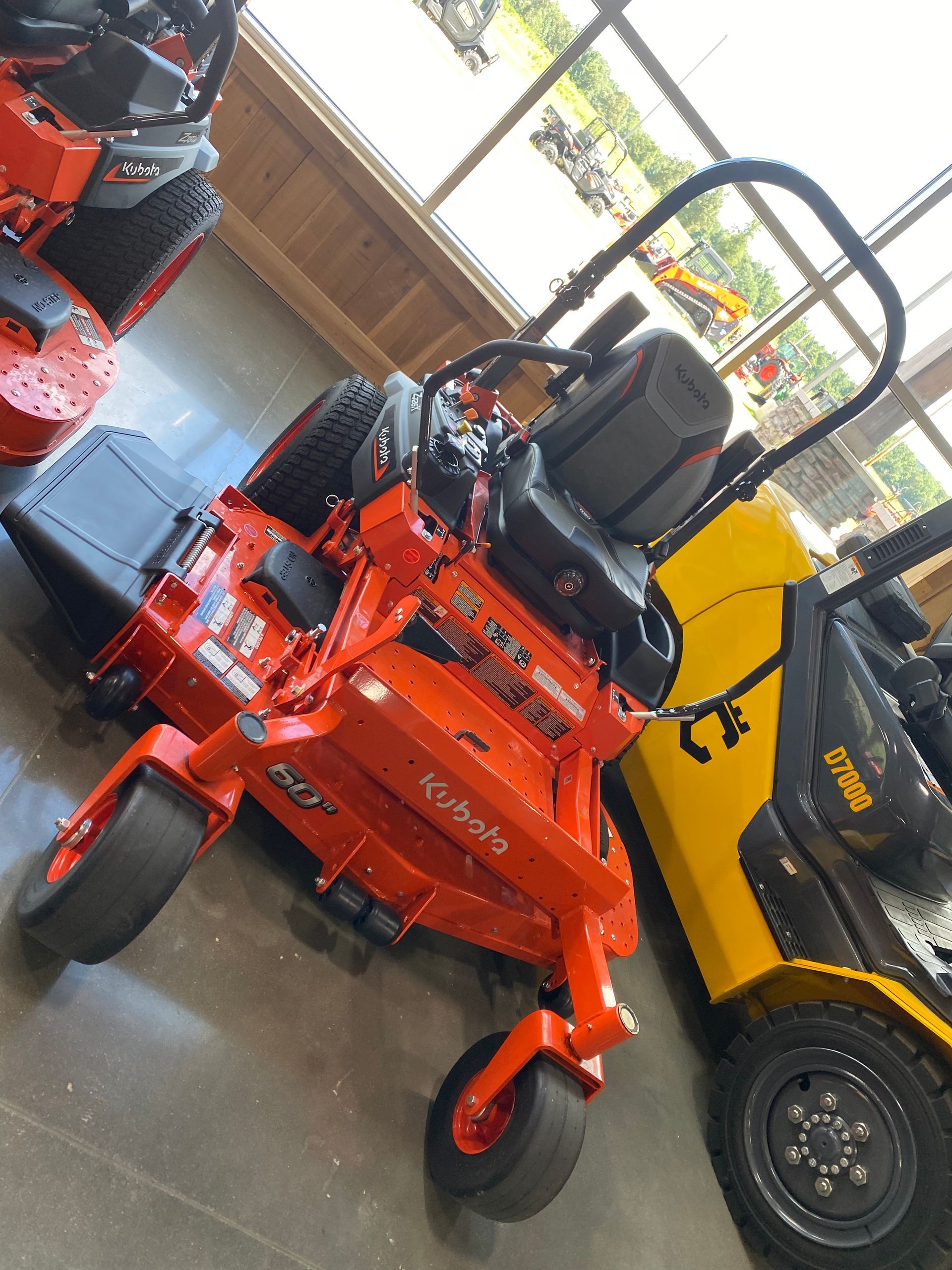 Orange Kubota zero-turn lawnmower, angled view, on display inside a showroom next to a yellow forklift.