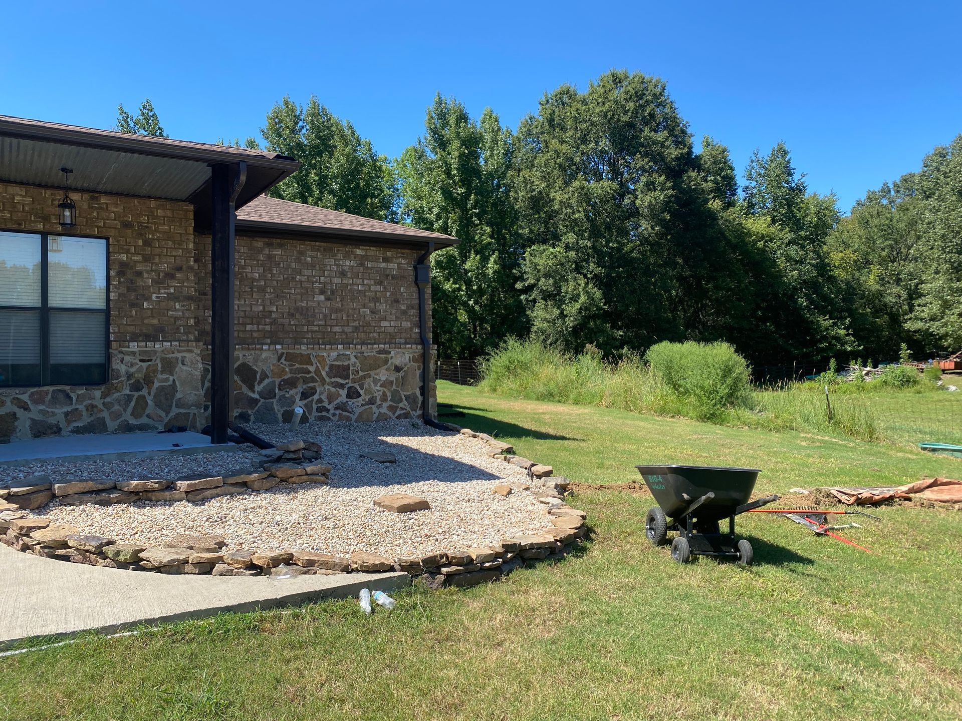 Exterior of a stone-walled building with gravel landscaping, a lawn, and a garden cart on a sunny day. Trees in the background.