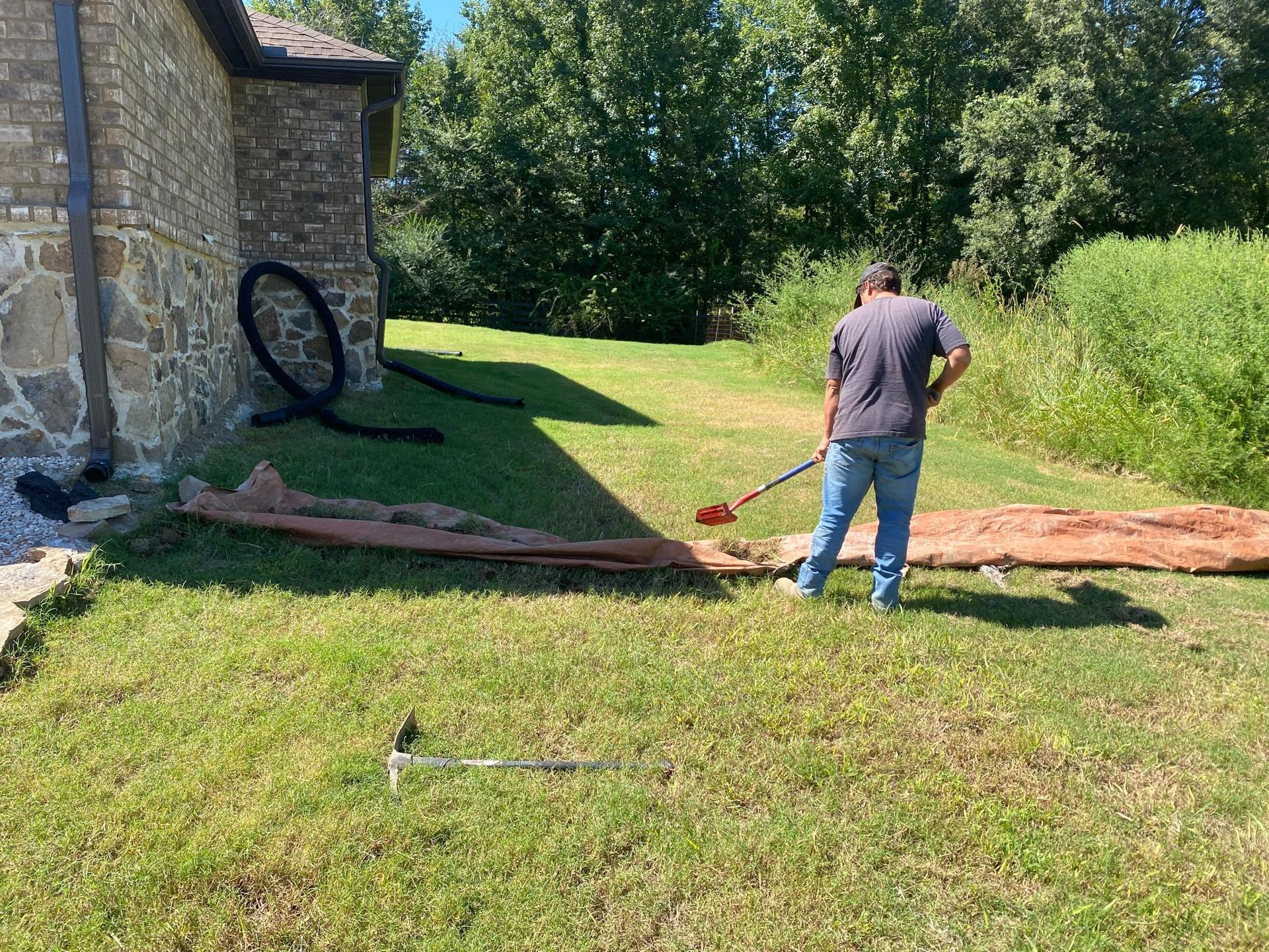 Man using a shovel to dig in a grassy yard next to a house. A long piece of reddish material is on the ground.