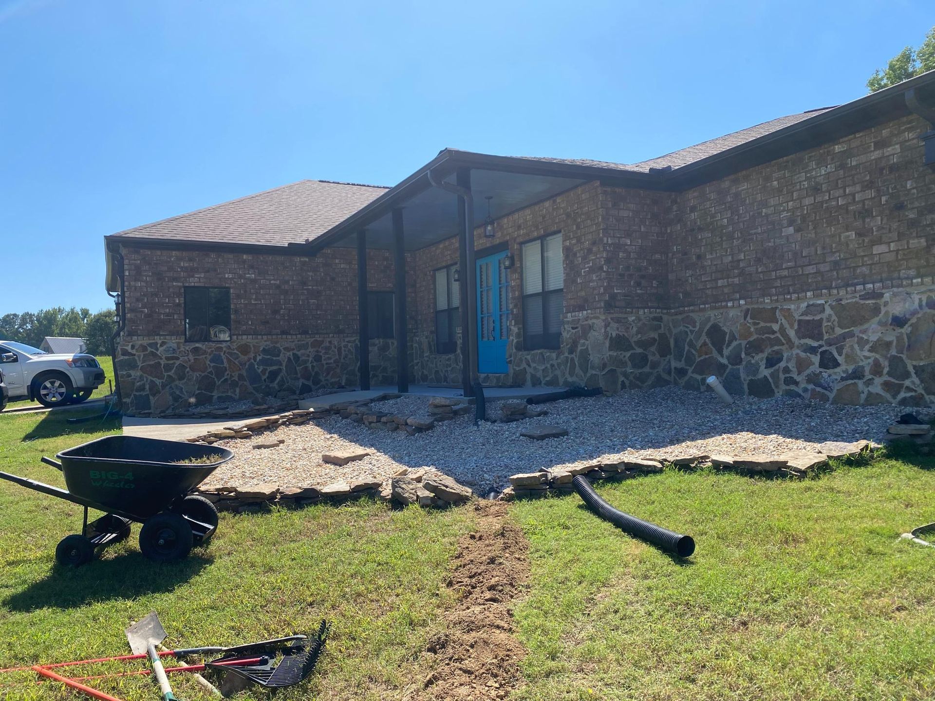 Exterior view of a brick and stone house undergoing landscaping. A wheelbarrow, tools, and drainage pipe are visible in the yard.