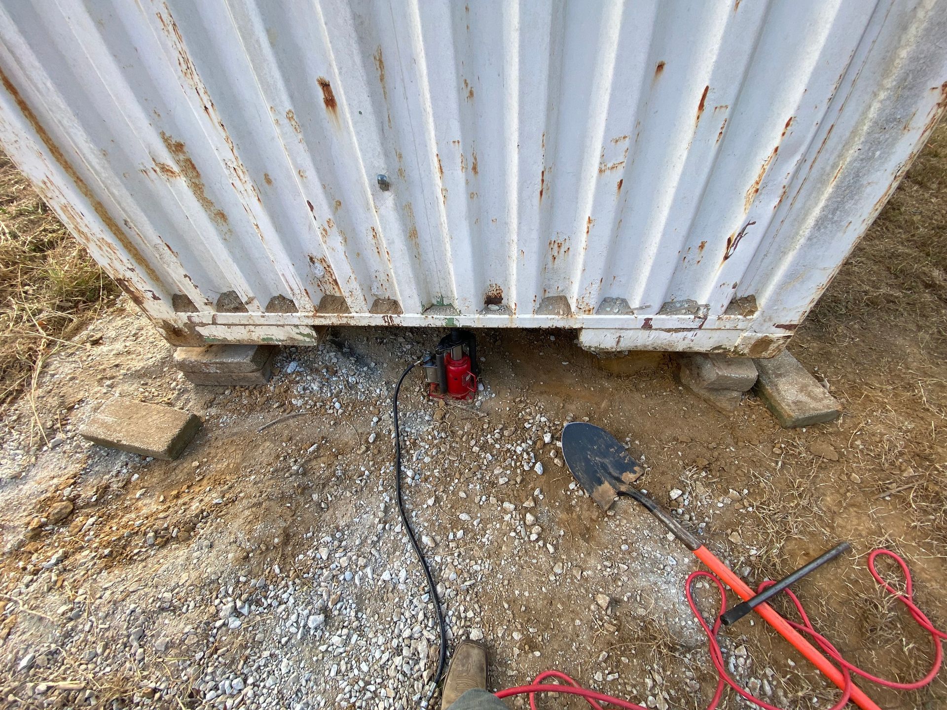 A shipping container is elevated with a jack, resting on concrete blocks in a dirt and gravel setting. A shovel and other tools are nearby.