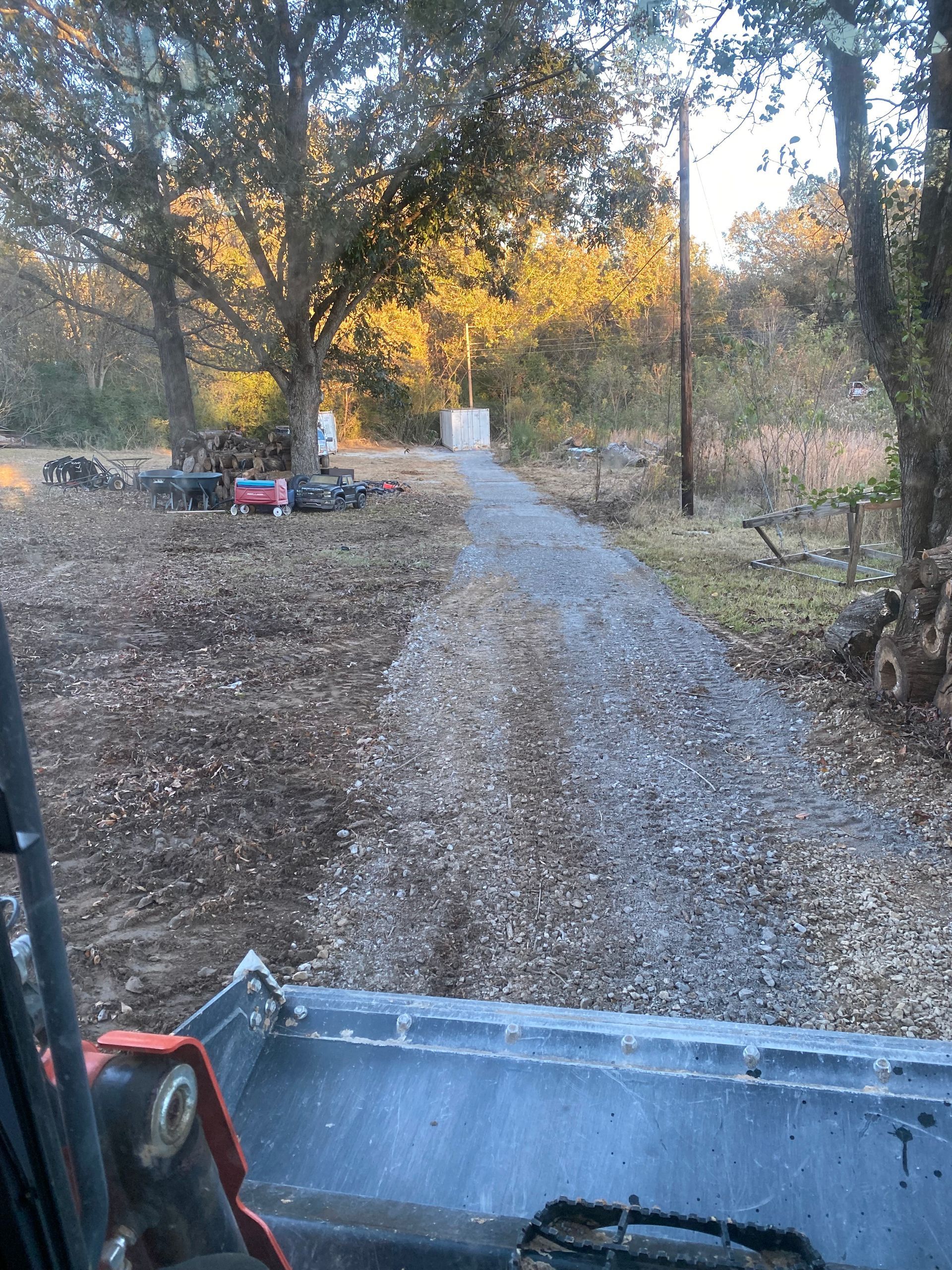 A gravel driveway being worked on by a loader with a rural setting. Bright sunlight illuminates the trees in the background.