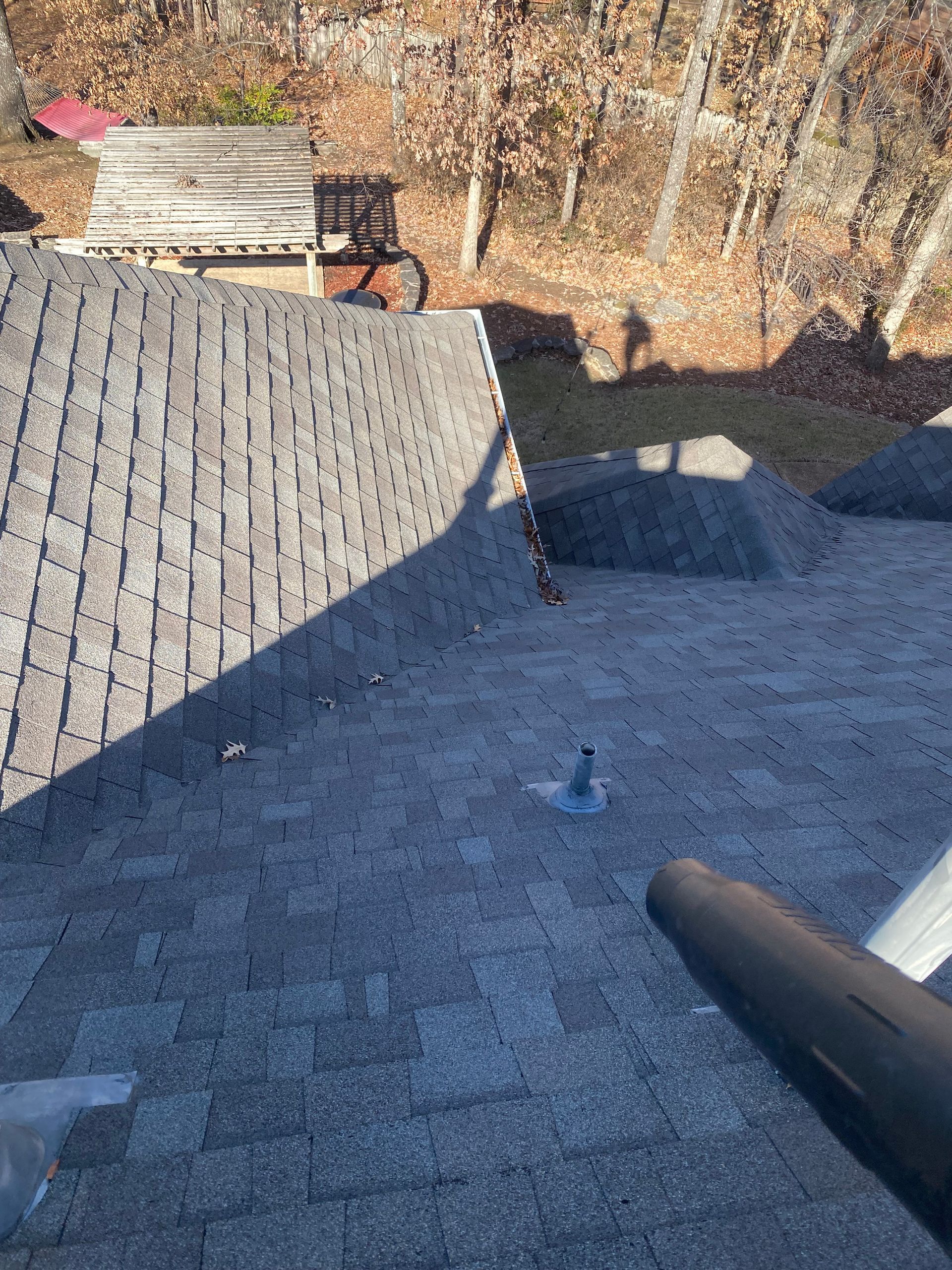 A person using a leaf blower to clean debris from a gray asphalt shingle roof on a sunny day.