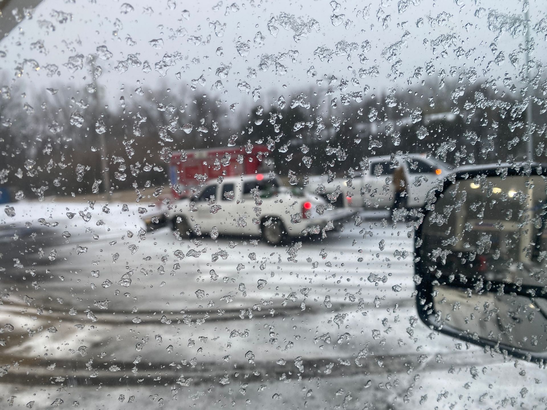 Snow-covered scene with a car window covered in ice. Two white pickup trucks are parked nearby.