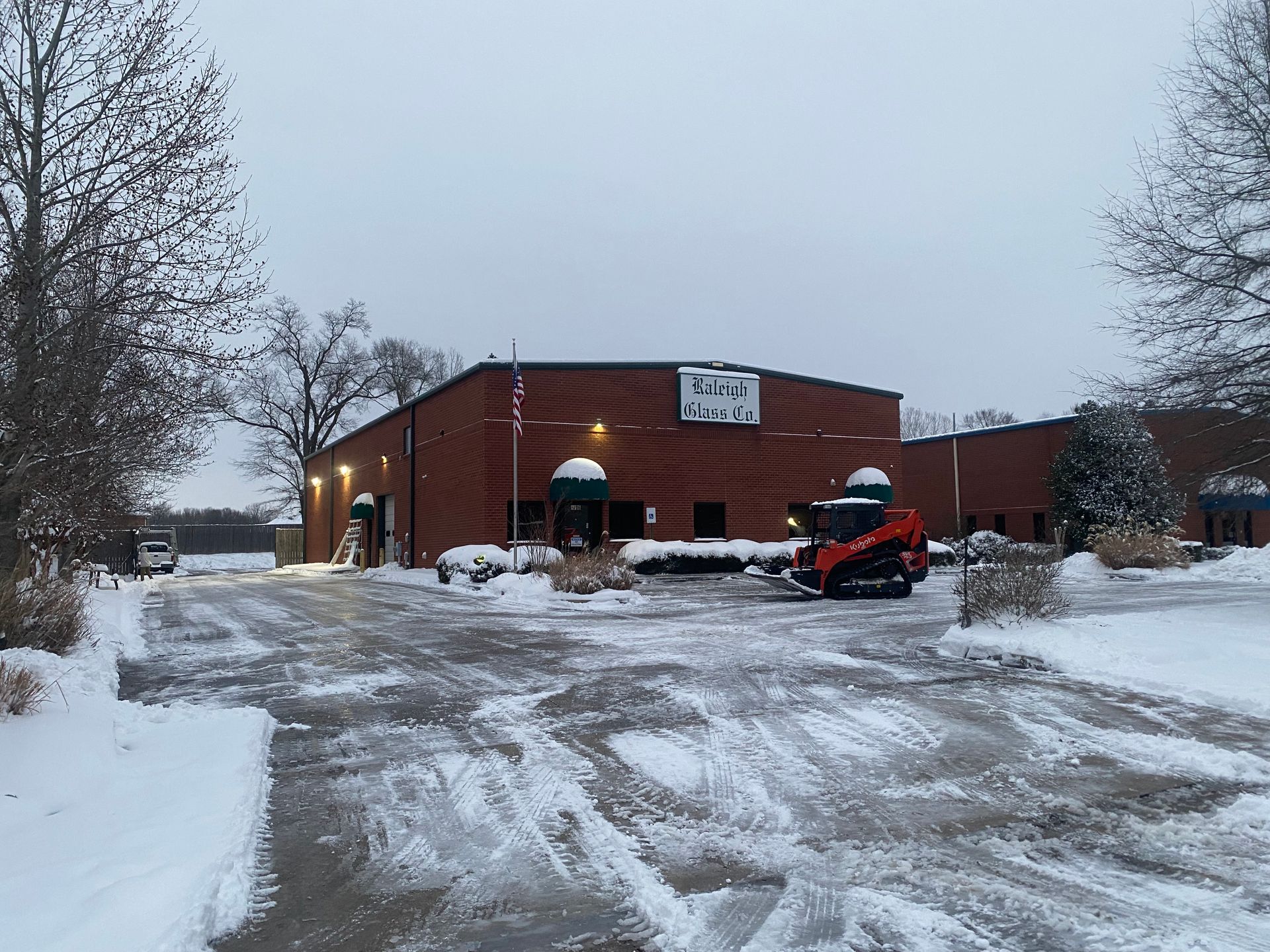 Snowy scene of a brick building with a small skid steer in front. The building has a sign that is partially visible.