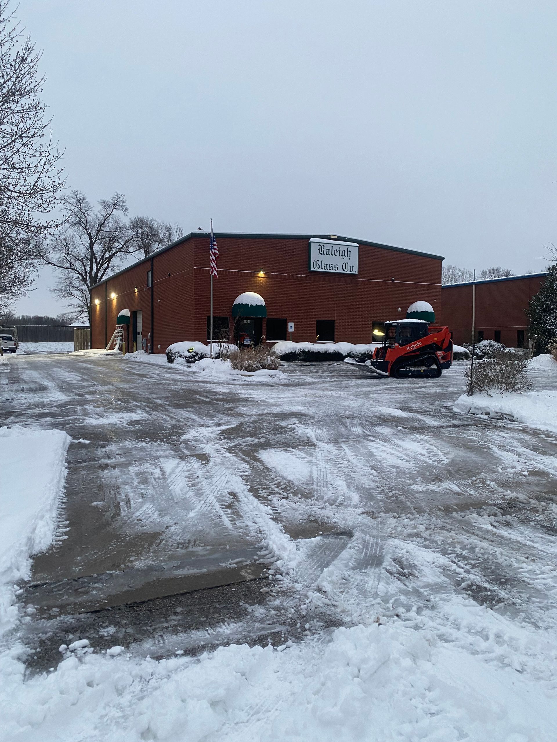 Snow-covered building with a small bulldozer clearing the parking area. Overcast, winter scene.