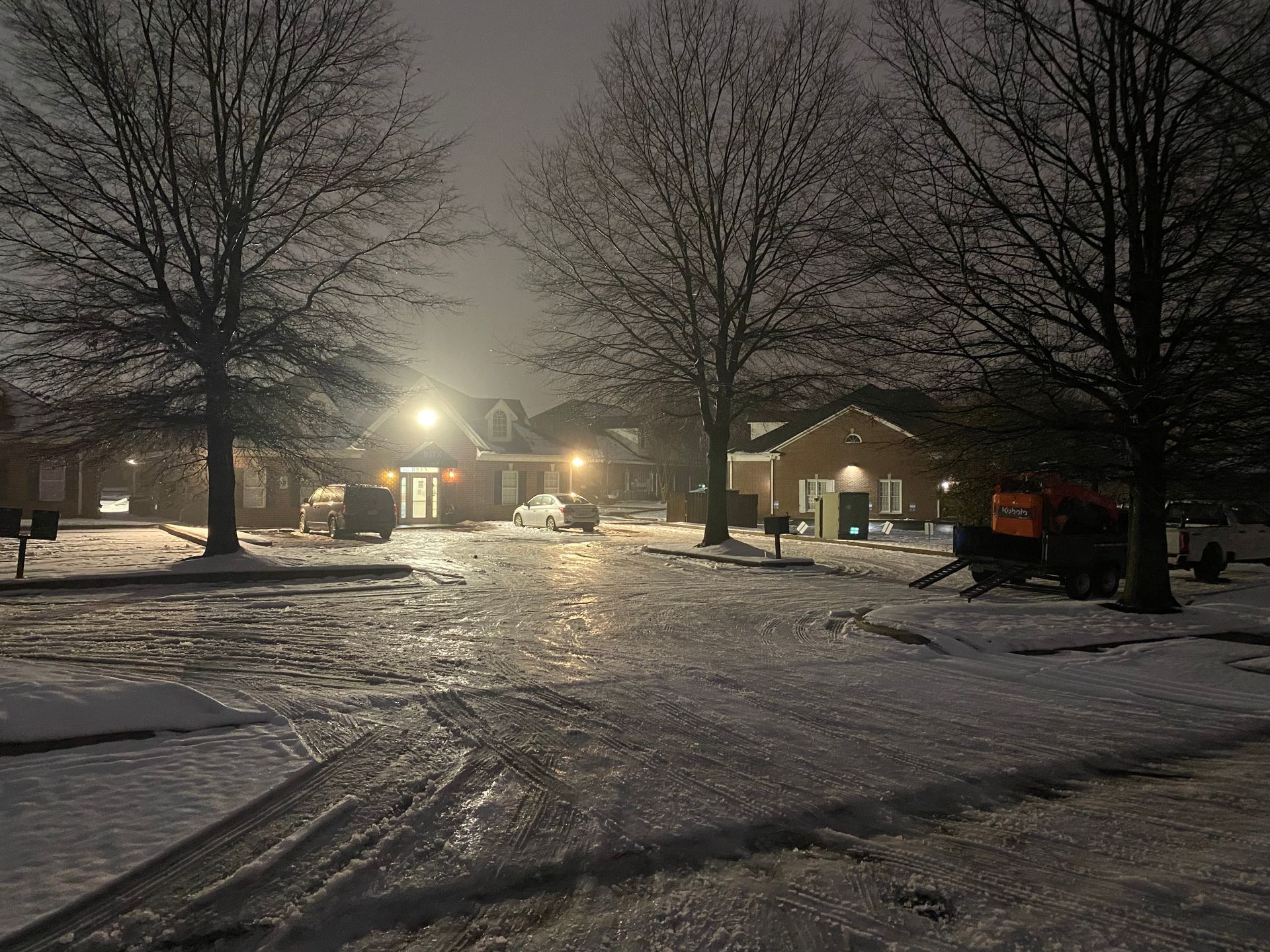 A snowy neighborhood street at night. Buildings and bare trees are illuminated by streetlights.