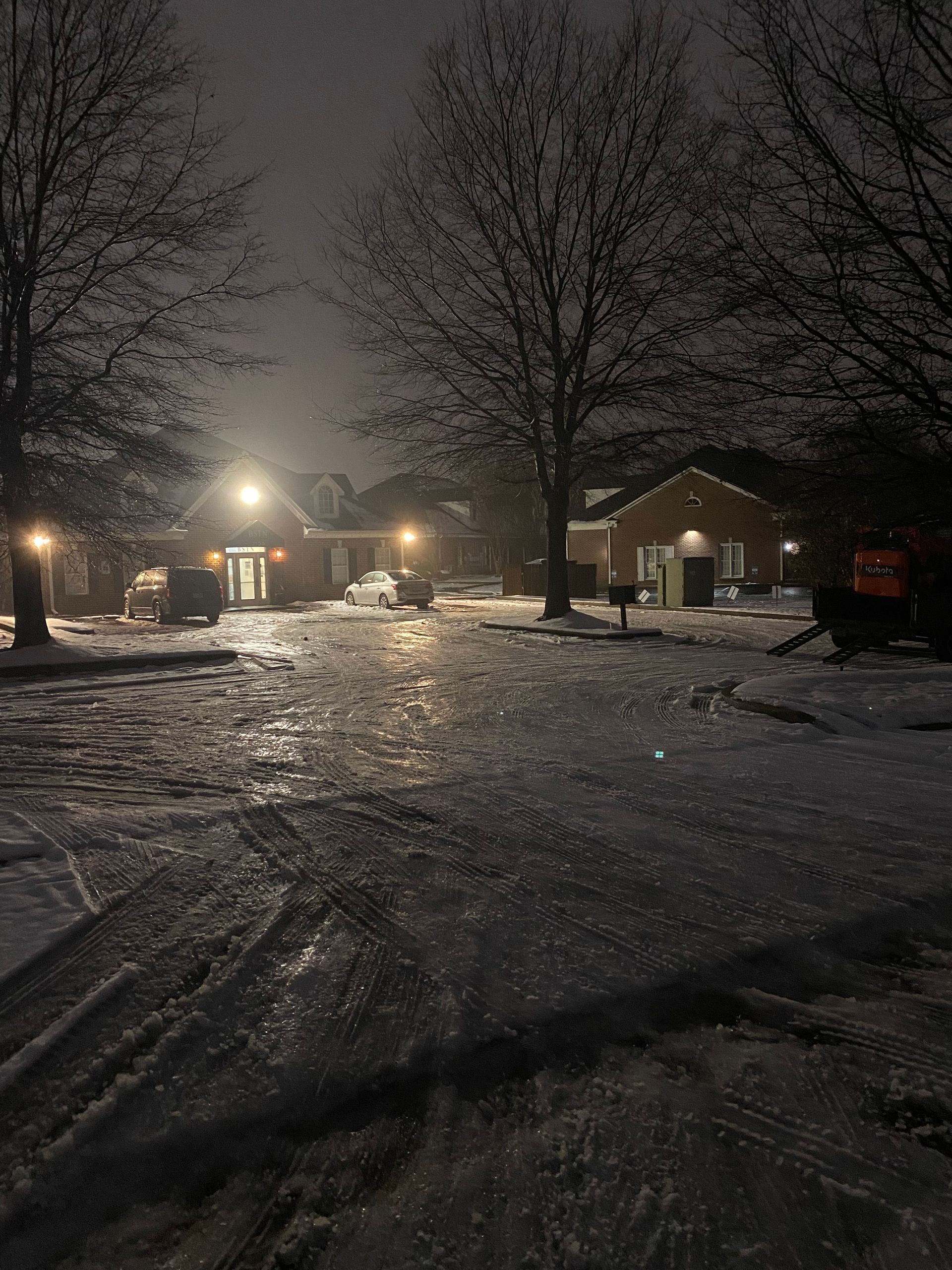 Snowy nighttime scene of a residential street with houses and bare trees illuminated by streetlights and building lights.