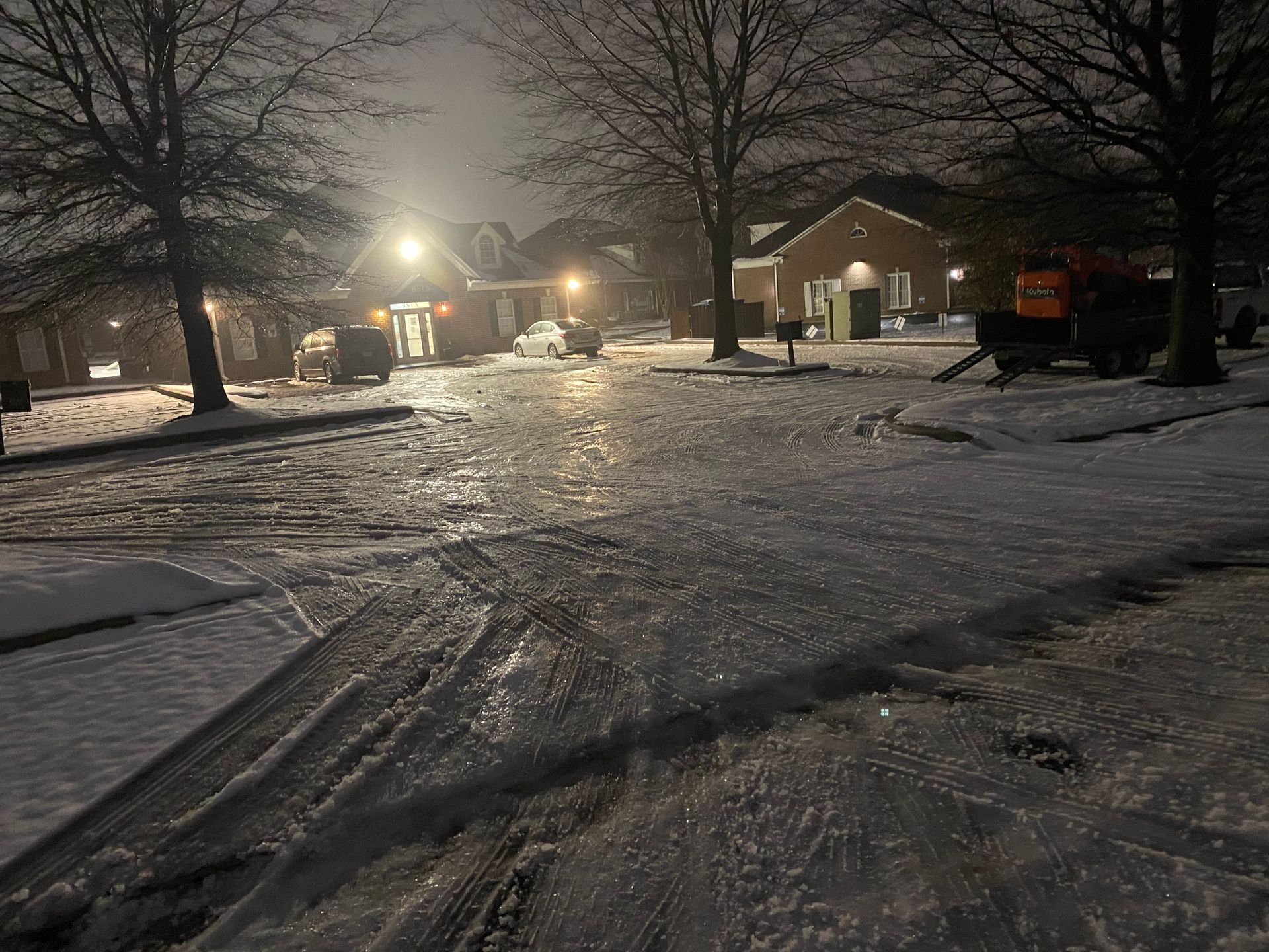 Snow-covered neighborhood at night with icy roads. Lights from houses and streetlamps illuminate the scene.