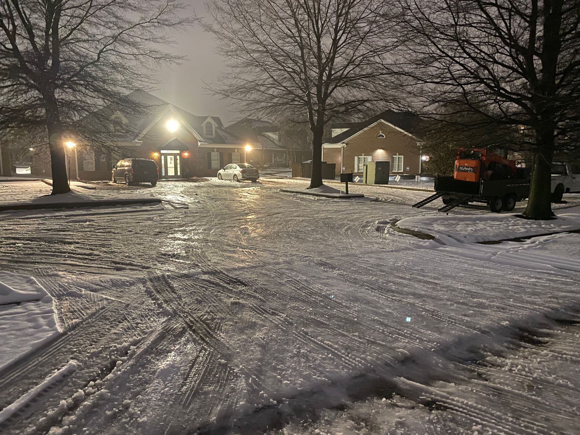 Snowy parking lot at night with buildings in the background. The ground is covered in ice and snow with tire tracks visible.