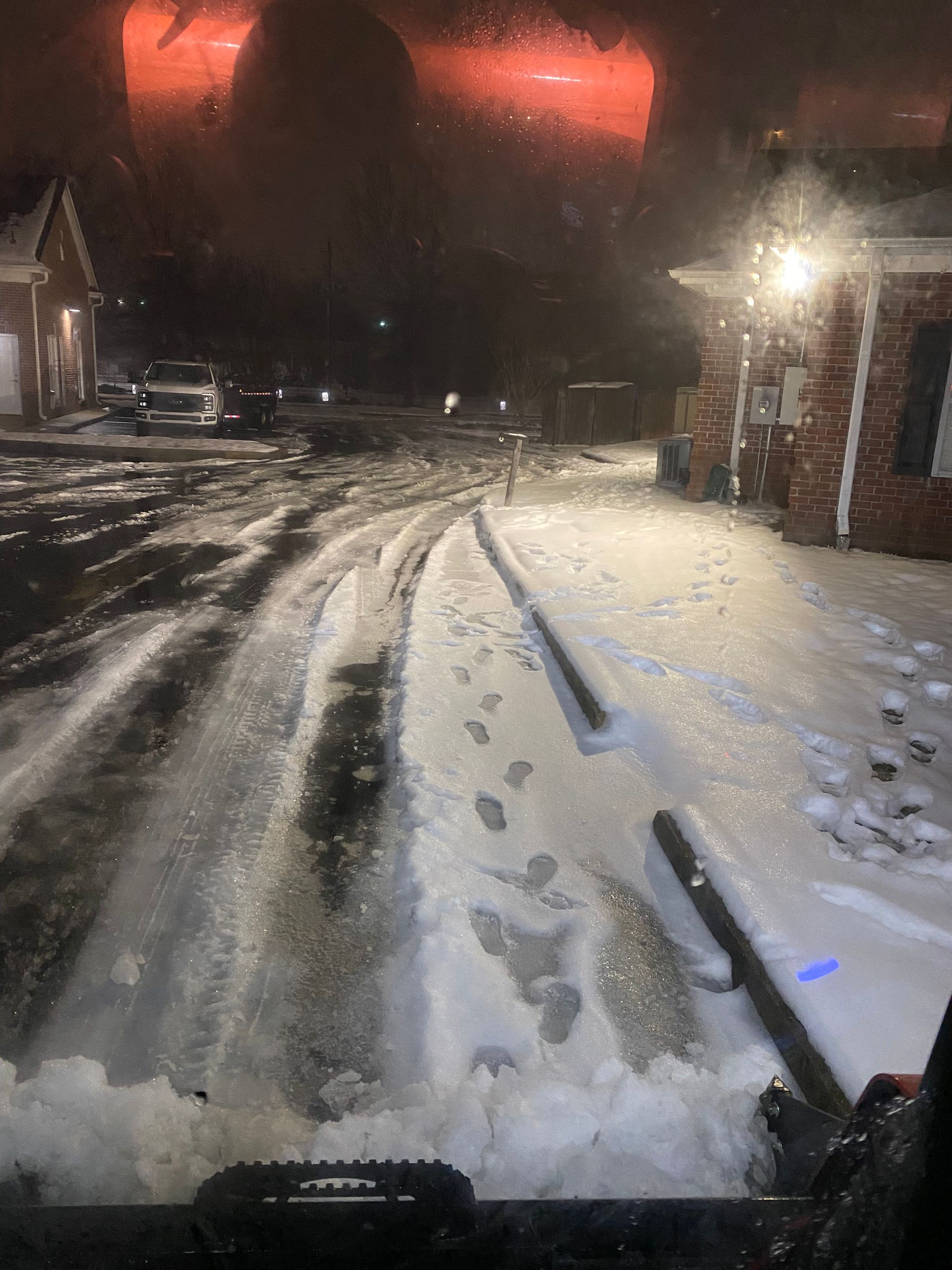 Snow-covered street at night, red sky above. Tracks cut through the snow, possibly cleared by a vehicle. Buildings line the sides.
