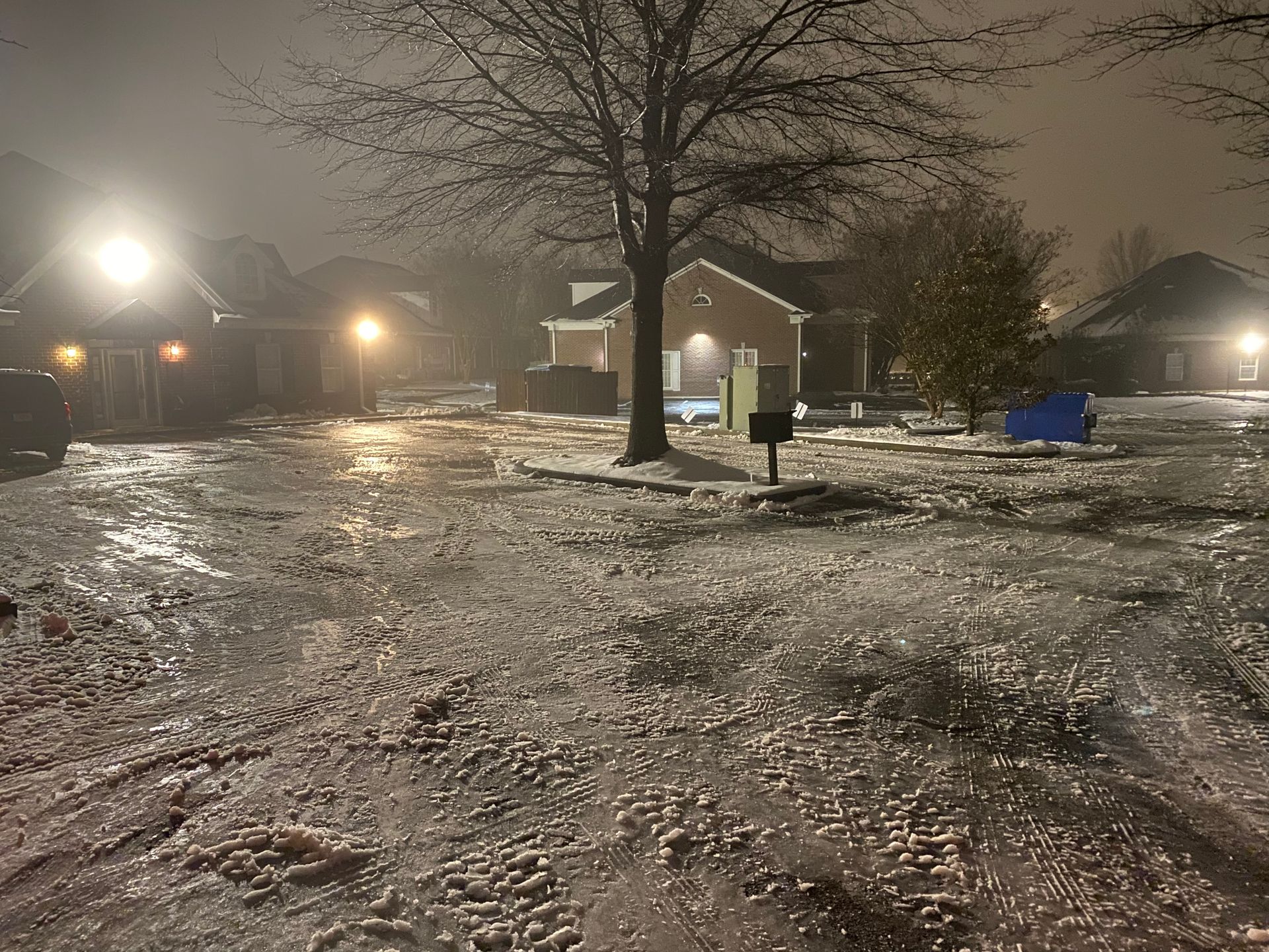 A snowy residential street at night with icy ground. Streetlights illuminate houses and a bare tree.