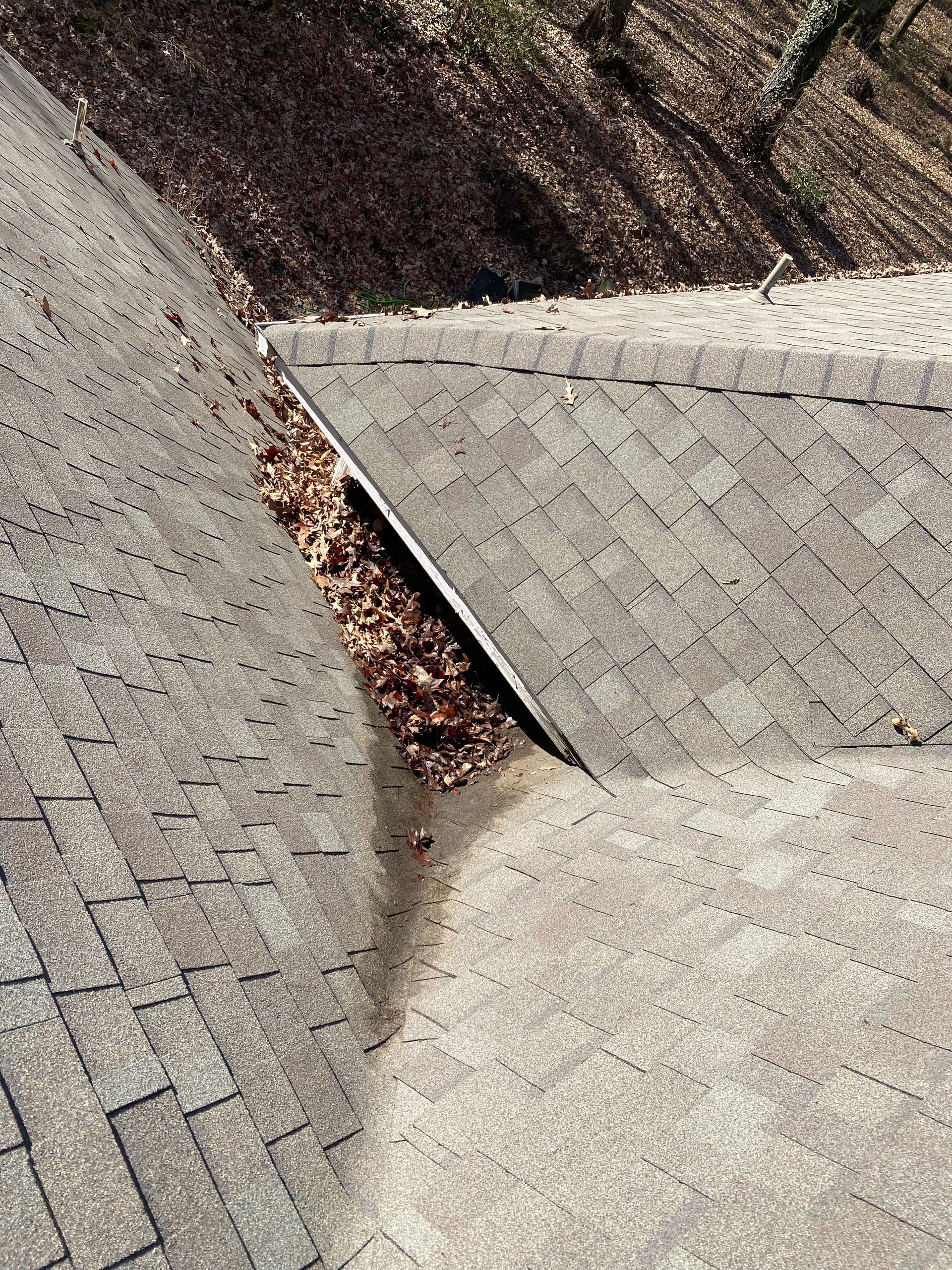 Overhead view of a roof with asphalt shingles, featuring a clogged valley with fallen leaves.