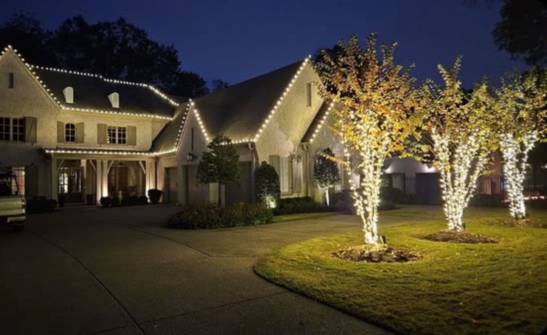 Night shot of a large house illuminated with white lights along the roof and wrapped around trees. A dark driveway leads to the house.