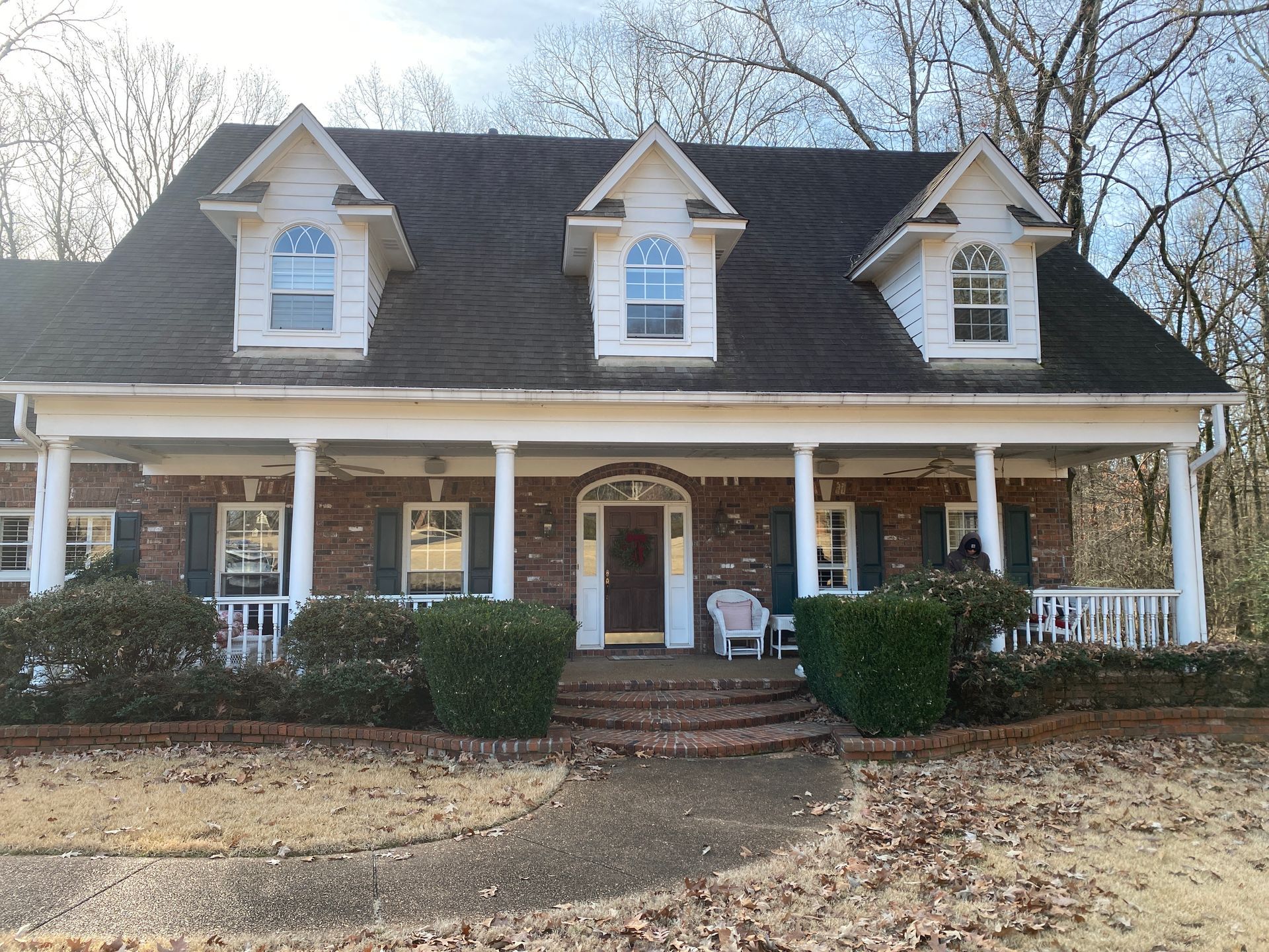 A brick house with a porch, three dormers, and a pathway. Bushes and a chair are on the porch, trees in the background.