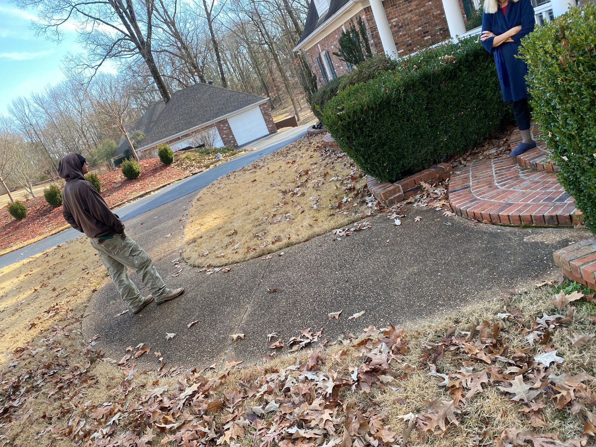 A person in a hoodie and camouflage pants stands on a speckled concrete path littered with brown leaves in front of a house.