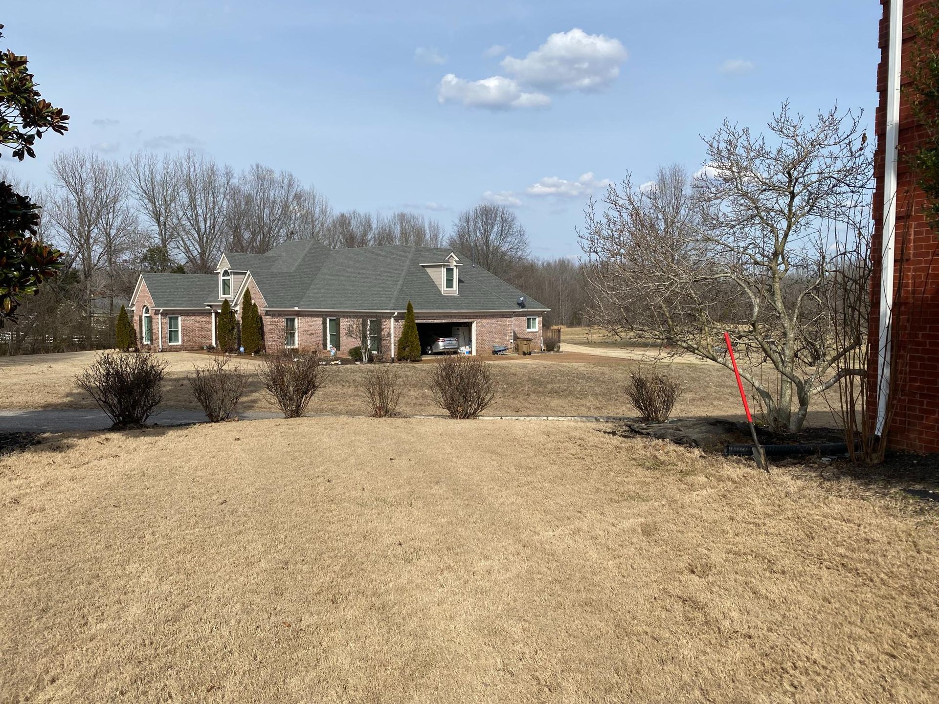 A single-story brick house with a dark green roof on a sunny day. Brown shrubs and dormant trees dot the lawn.