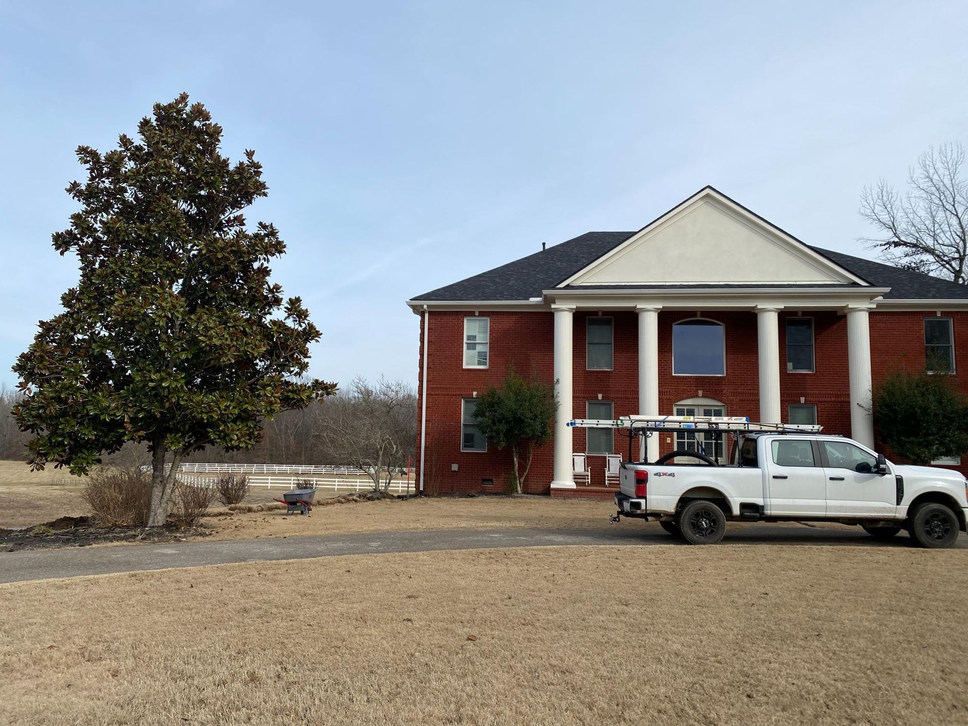 White truck parked in front of a large brick house with white columns. A tree stands to the left. The sky is clear and blue.