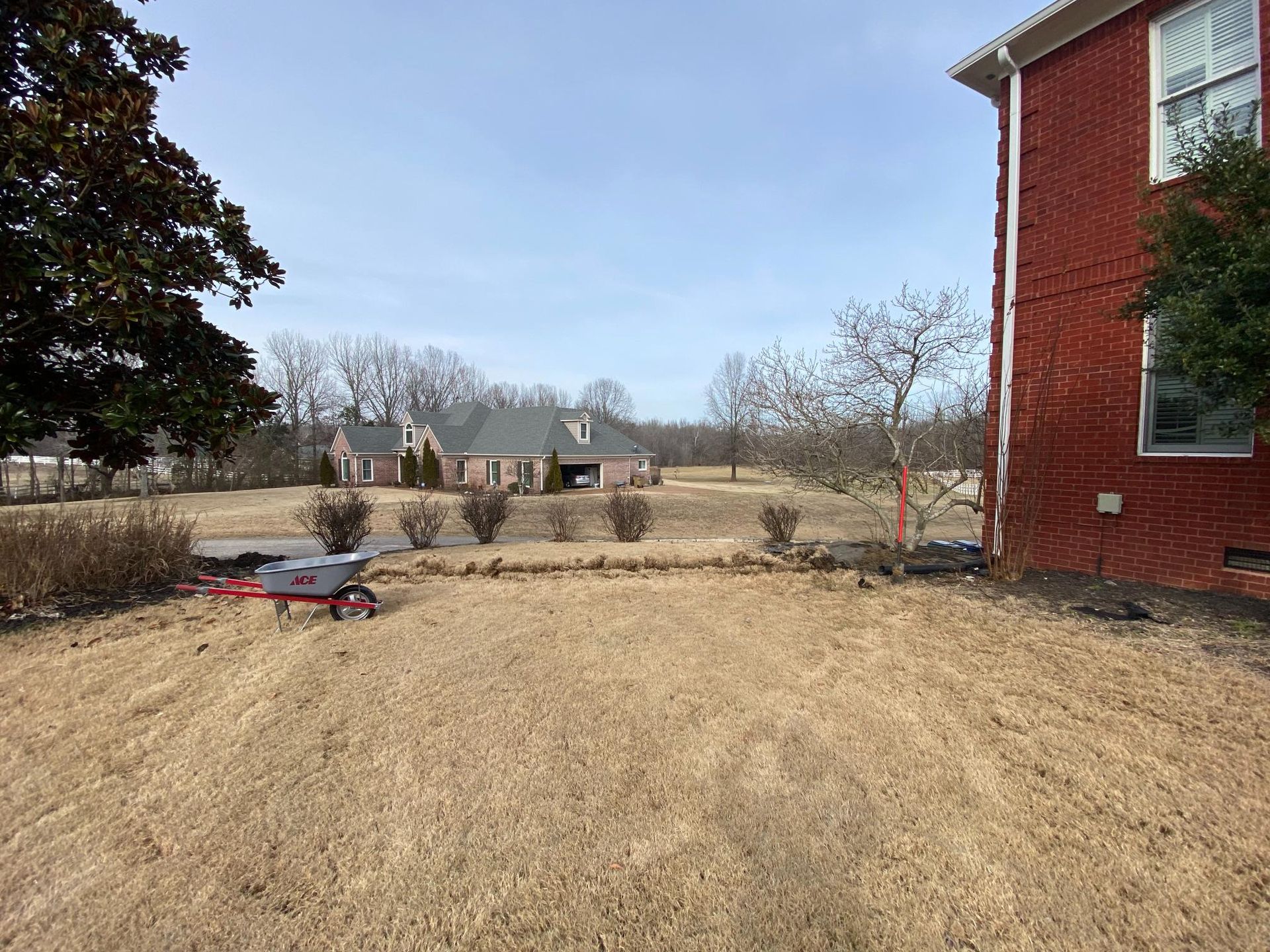 A red fertilizer spreader on a brown lawn near a brick building. Several houses are visible in the distance under a blue sky.
