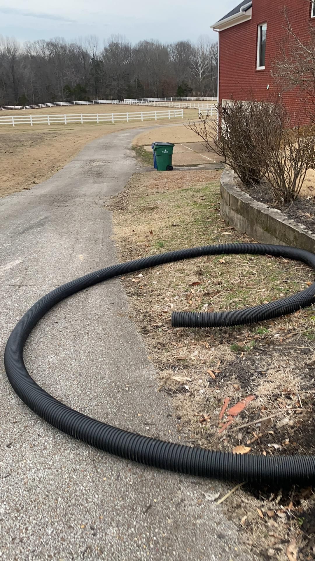 Black corrugated drain pipe coiled on a driveway next to a red building and green trash can. A fence and trees are in the background.