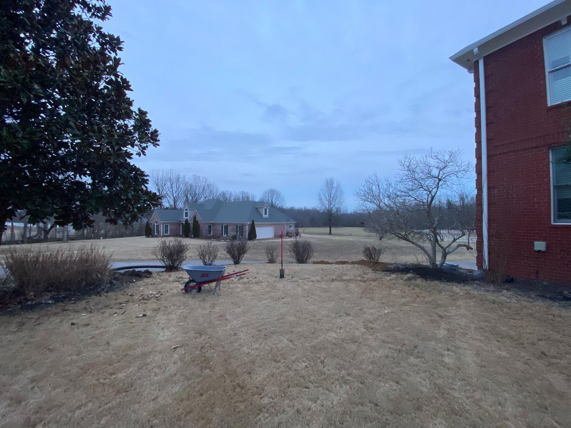Overcast backyard scene with a lawn, wheelbarrow, and red brick house on the right. Further in the distance, a house can be seen.