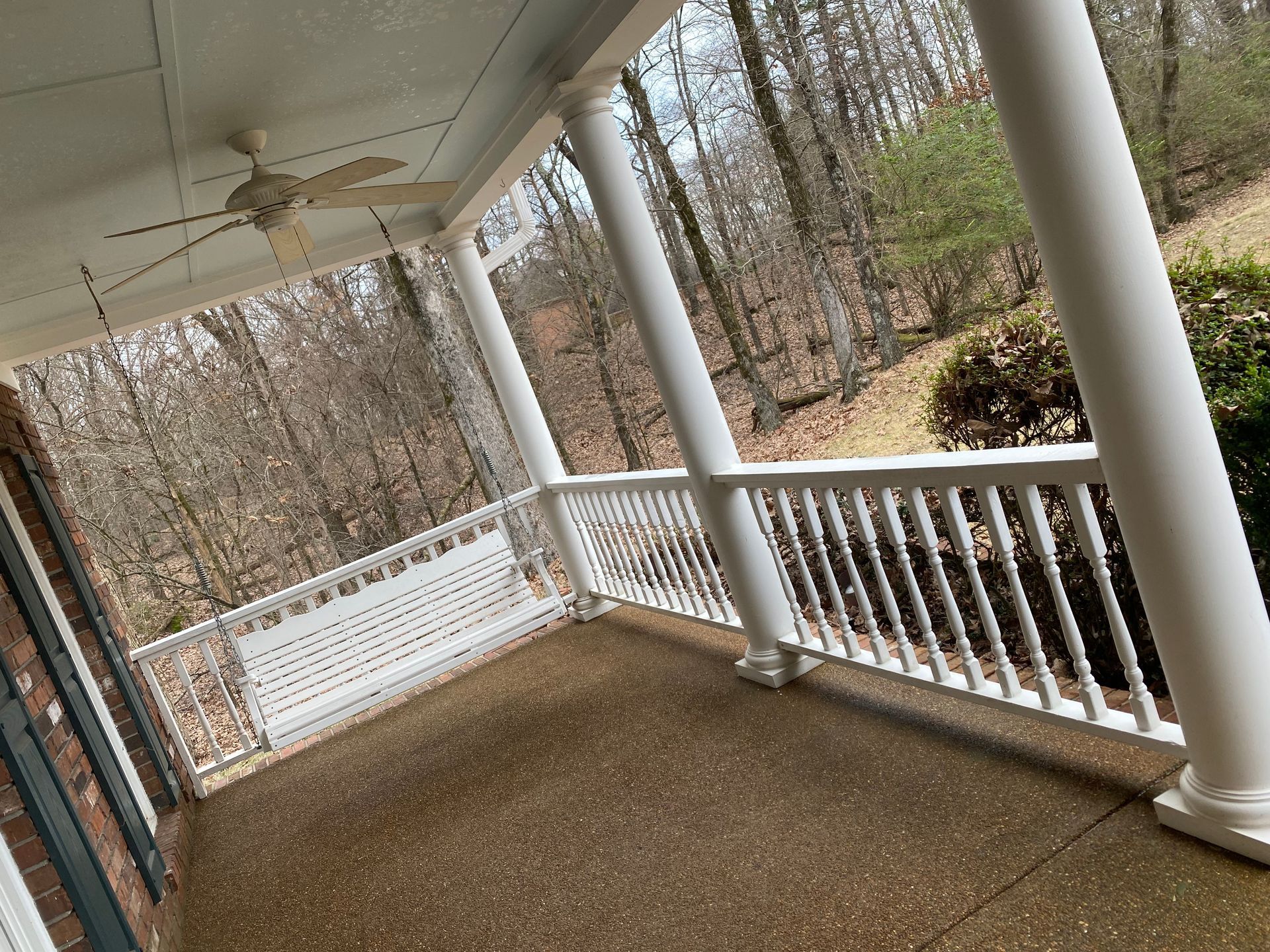 Covered porch with white columns and railing overlooking a wooded area. A porch swing hangs on the left.