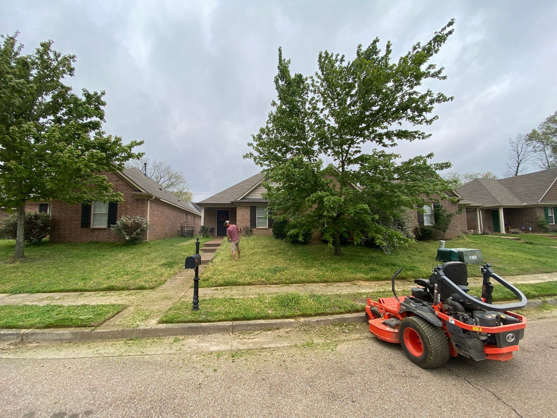 Lawn care in progress: A person mows a green lawn in front of a brick house with a riding mower on a cloudy day.