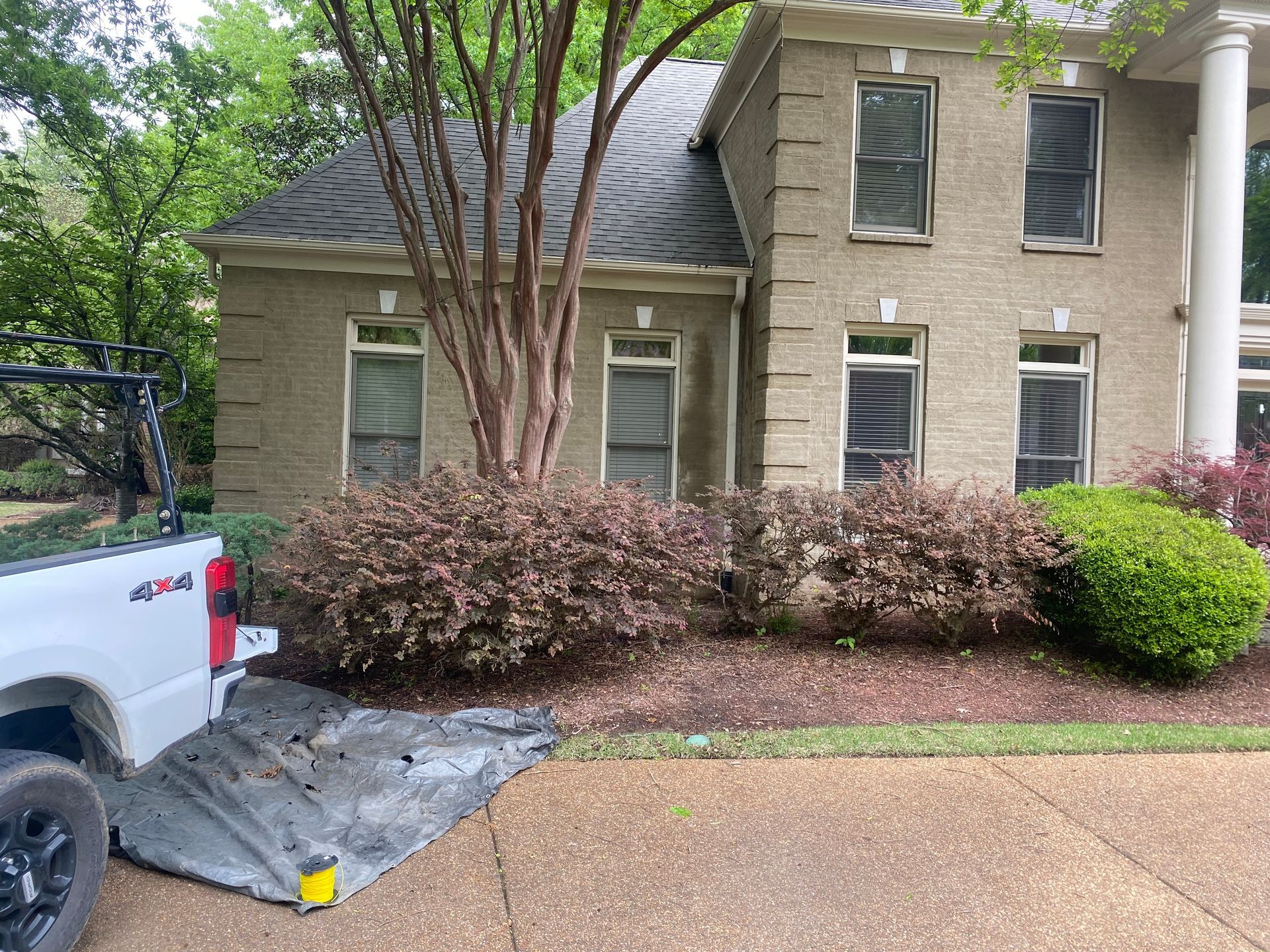 White truck parked on a driveway next to a house with dark roof, bushes, and tree. Grey ground cover and shrubs in front of house.