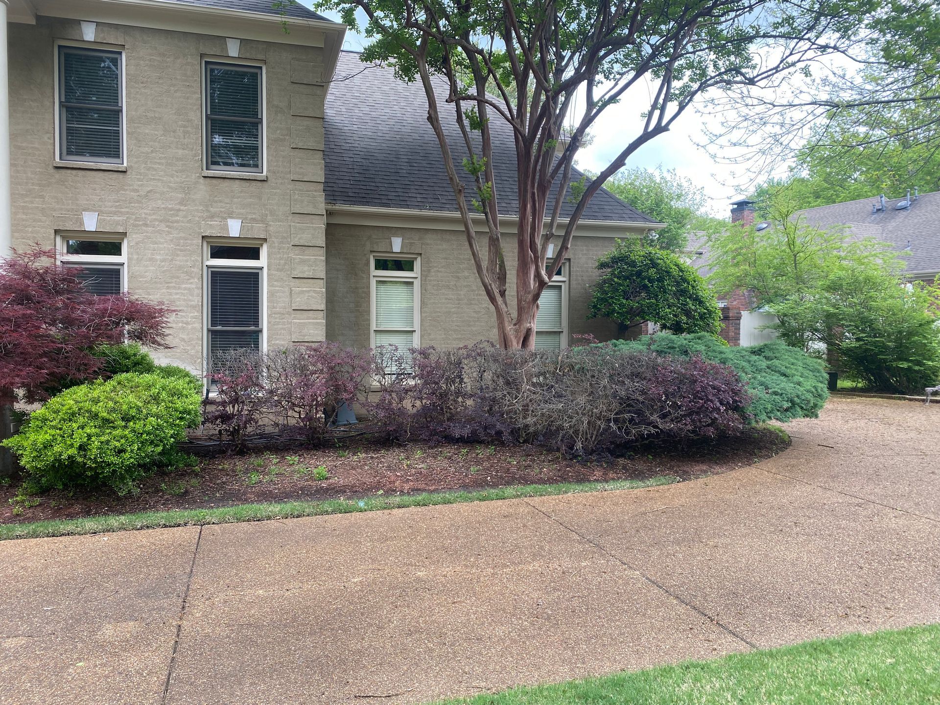 A two-story brick house with a gravel driveway, surrounded by colorful bushes and trees under a cloudy sky.