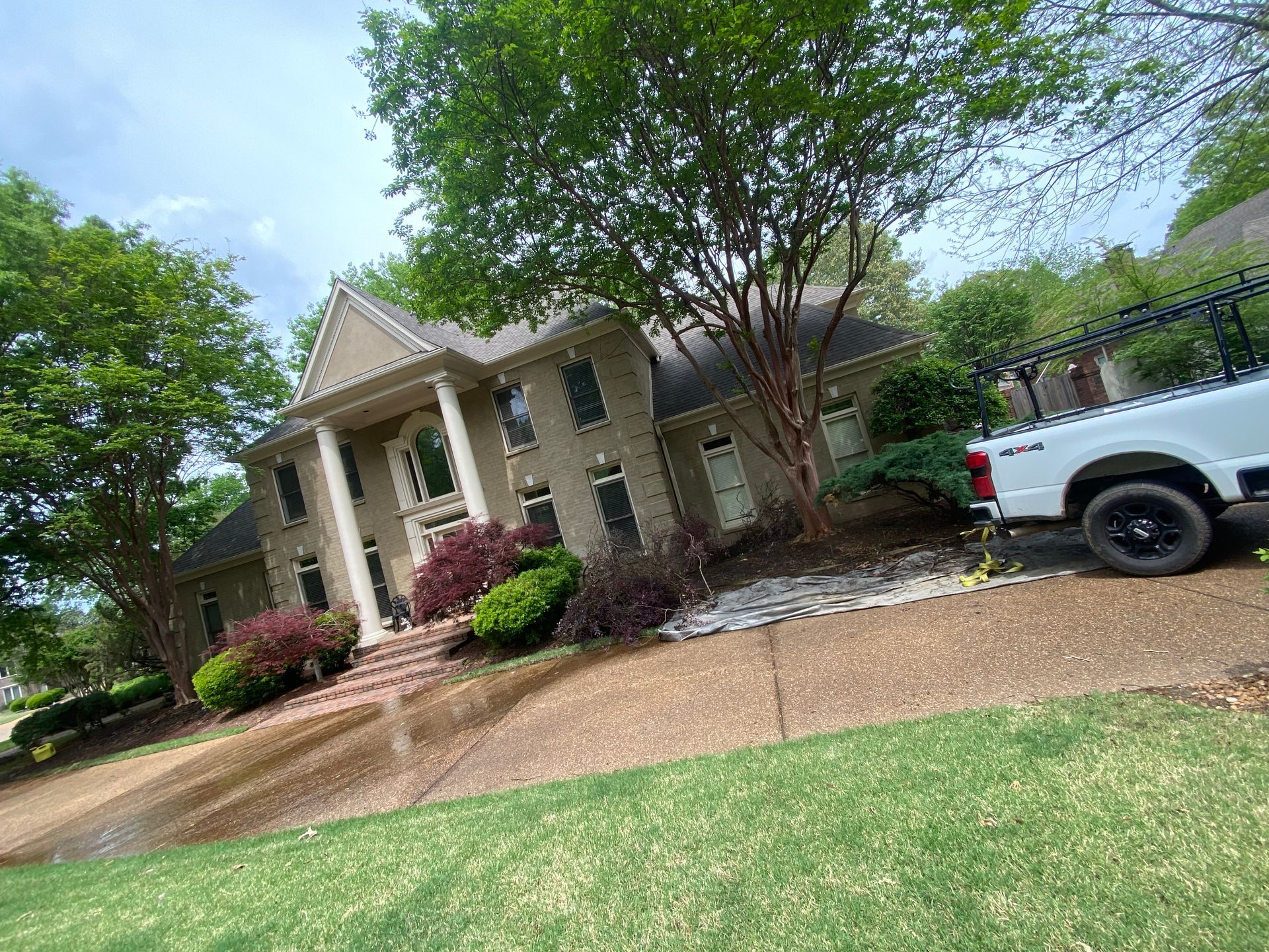Two-story beige house with large columns and a white truck with a ladder parked nearby. Lush green trees and bushes surround the property.