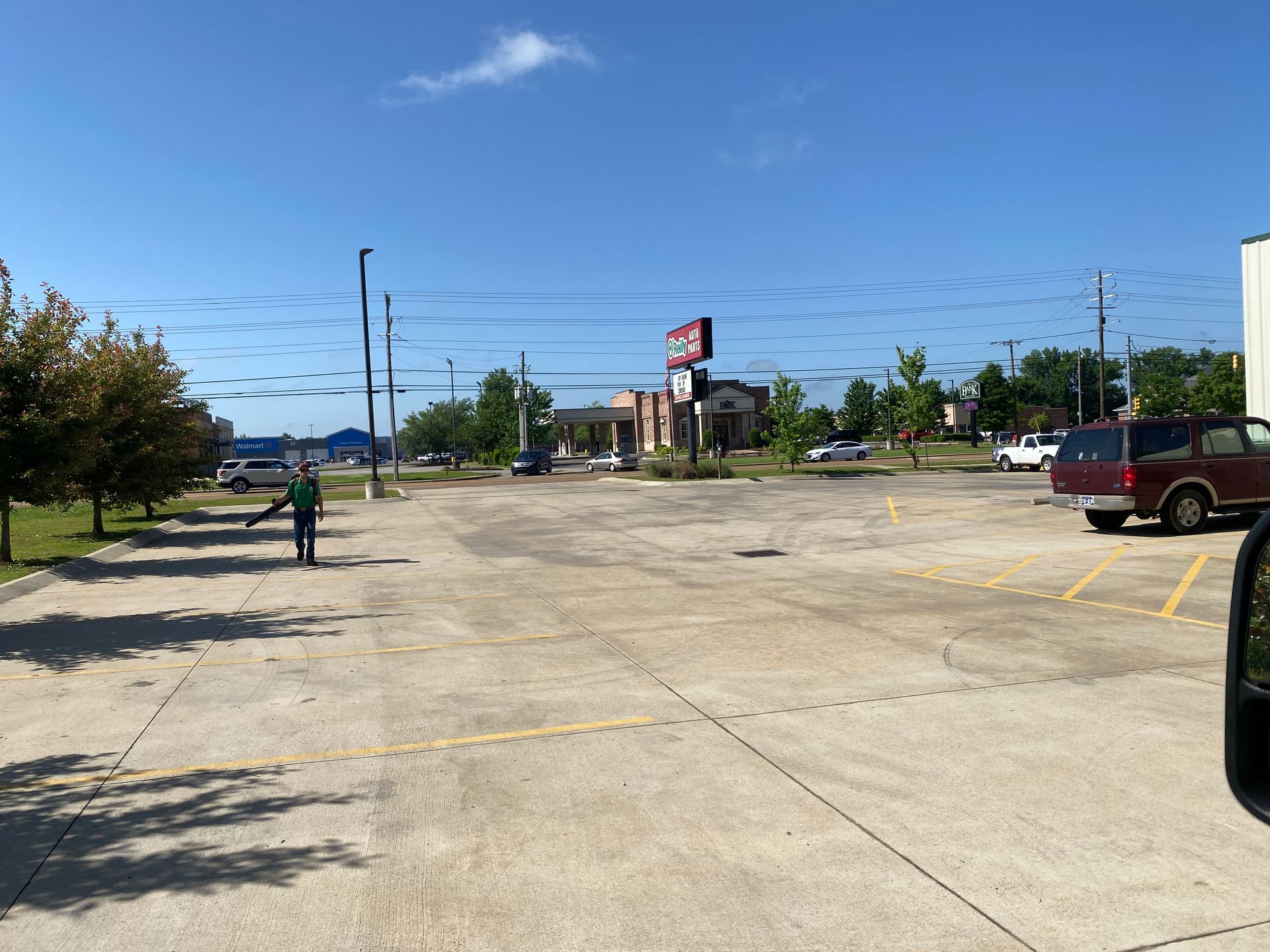 Person in green gesturing in a large, mostly empty parking lot on a sunny day, with buildings and a sign in the background.