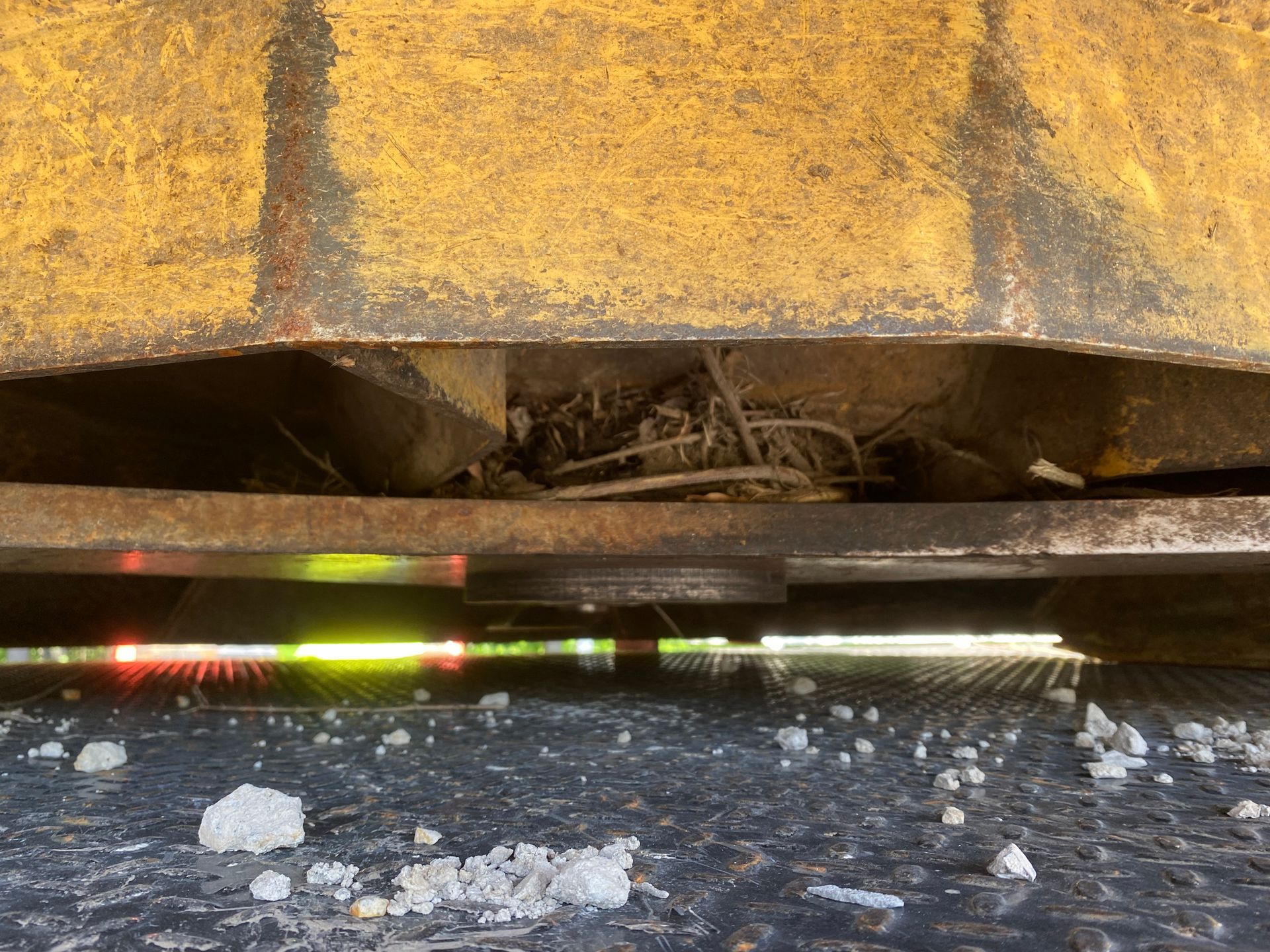 Underneath a yellow machine, debris rests between metal plates. Sunlight shines through the opening.