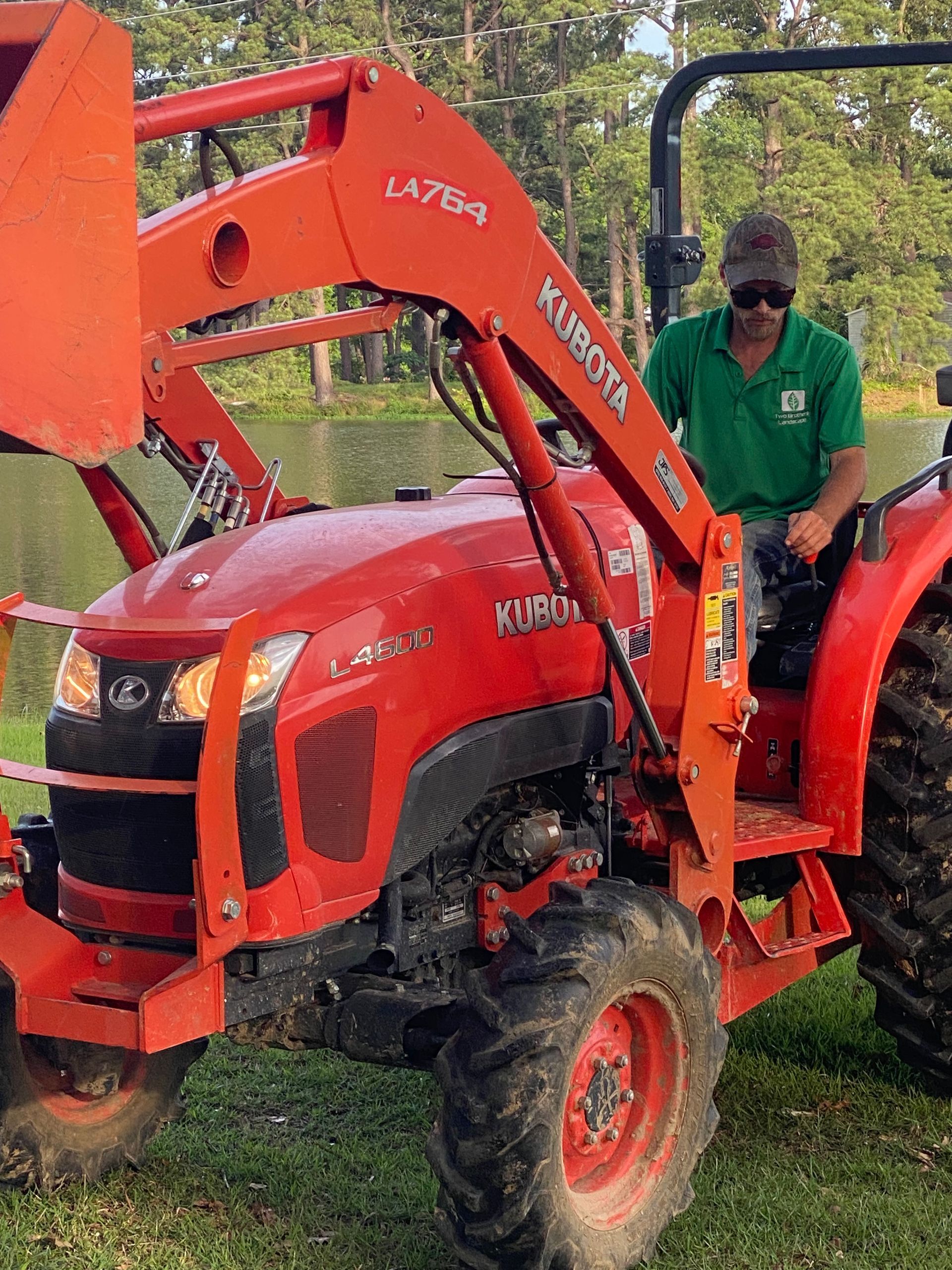 Man in green shirt operating a red Kubota tractor with a front loader. He is outdoors near a pond.