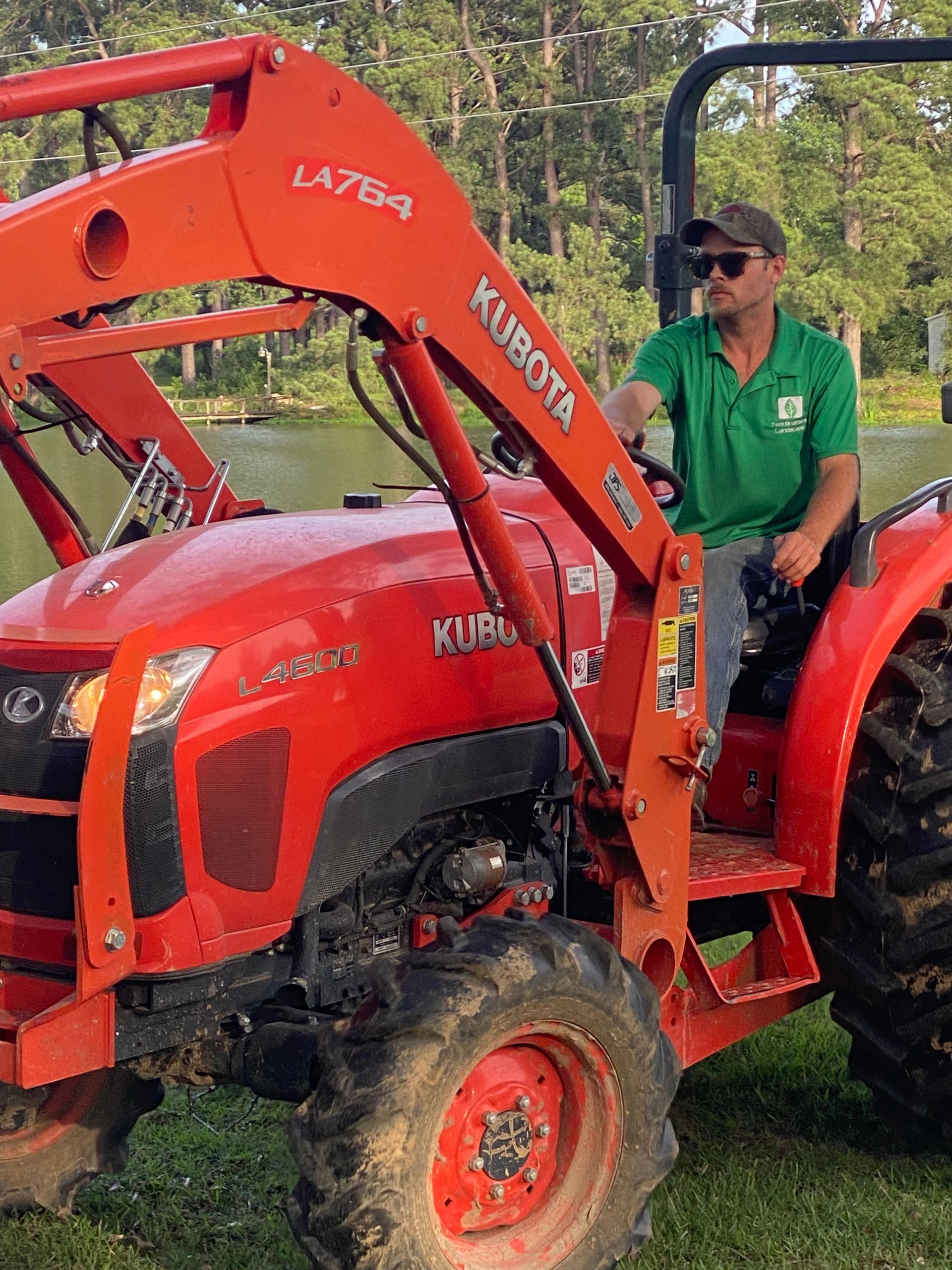 Man driving an orange Kubota tractor with a front loader outdoors near a pond, wearing a green shirt and a hat.