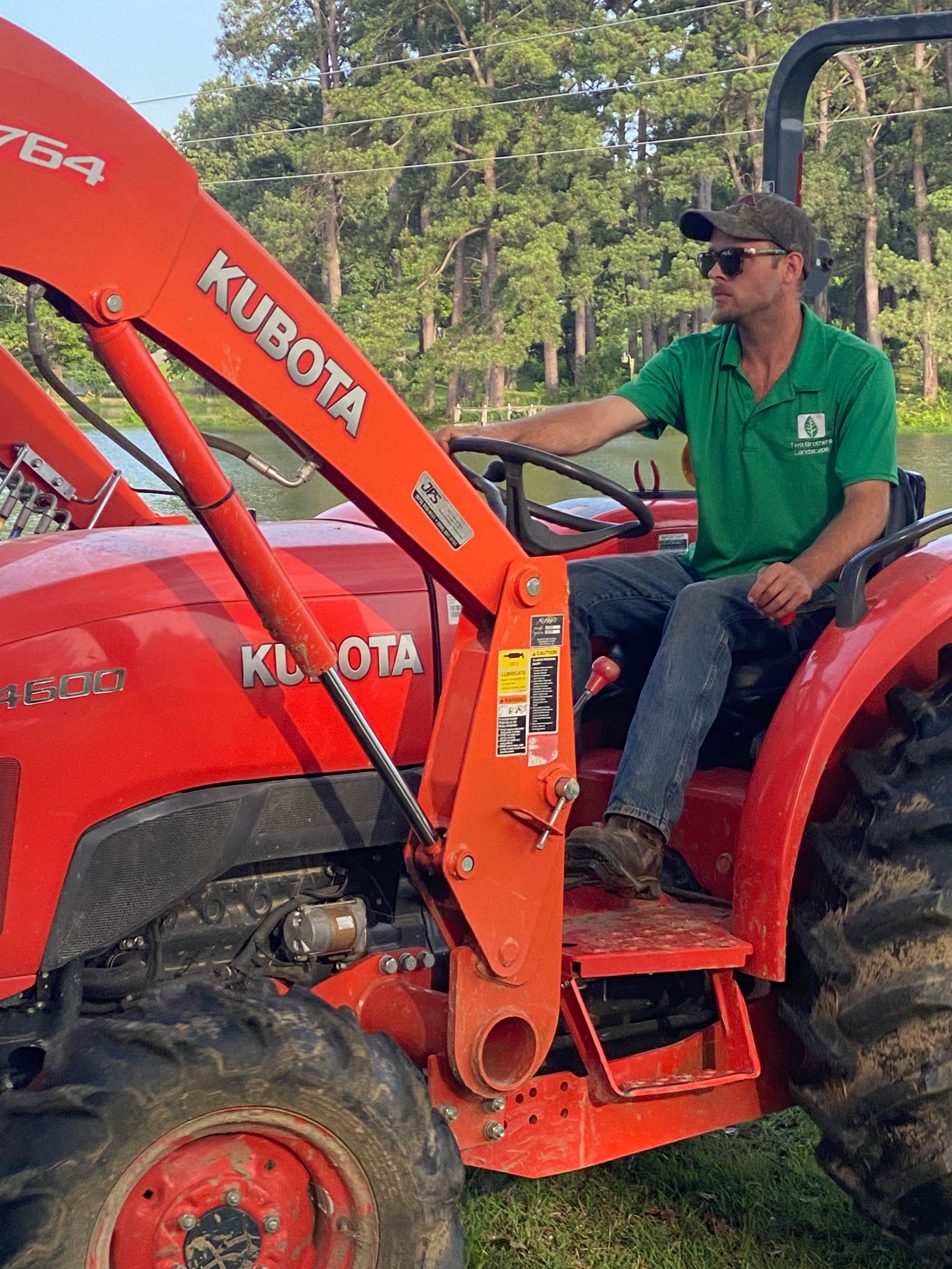 Man in green shirt and baseball cap driving an orange Kubota tractor outdoors.