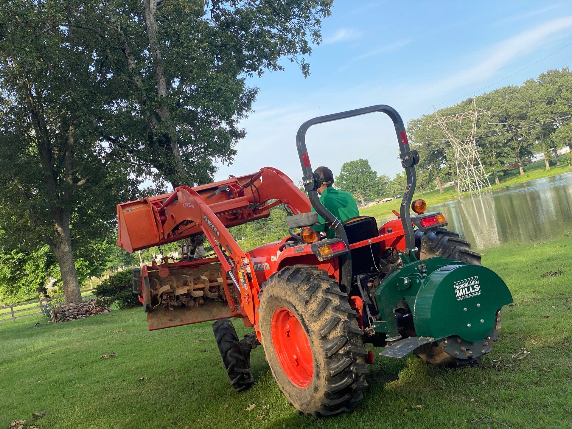 A person operates an orange tractor with attachments on a grassy field. The tractor is in front of a tree and near a body of water.
