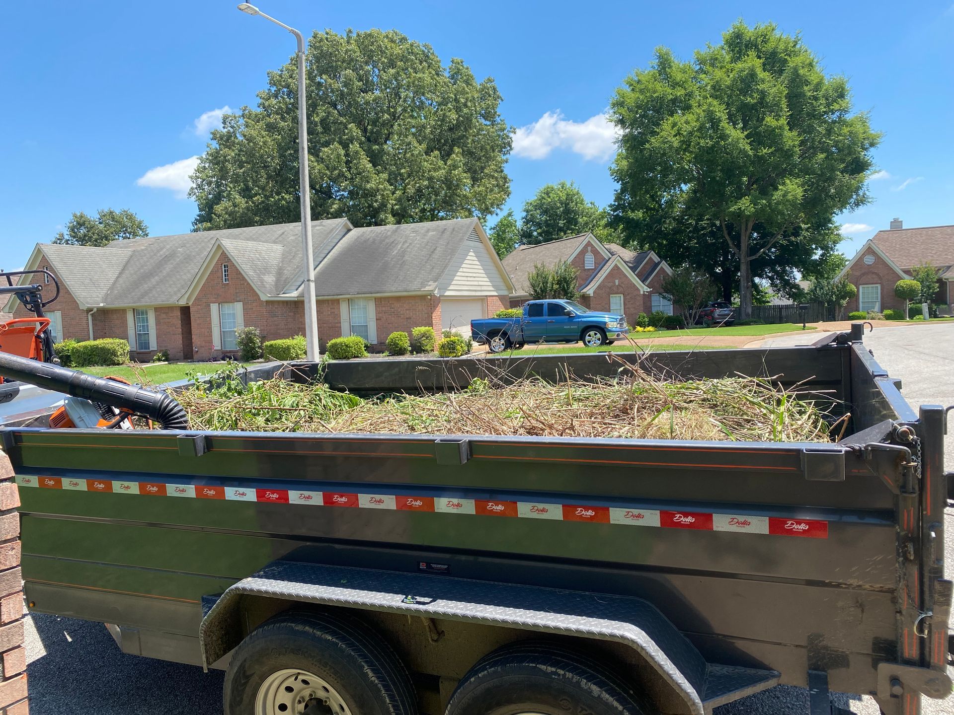 A trailer filled with yard waste sits in front of houses on a sunny day. A blue truck is parked nearby.