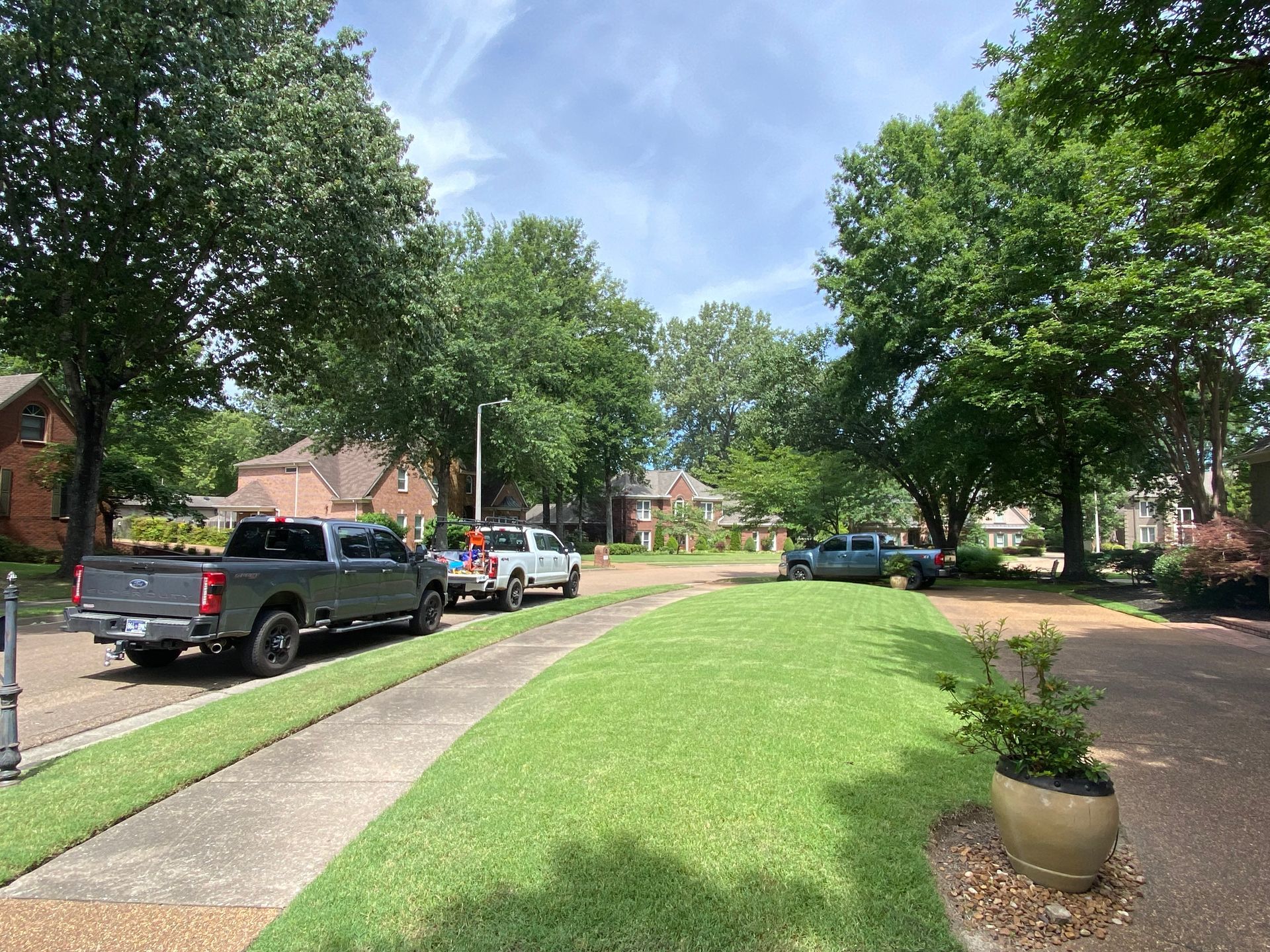 A residential street lined with trees, green lawns, and several parked pickup trucks under a partly cloudy sky.