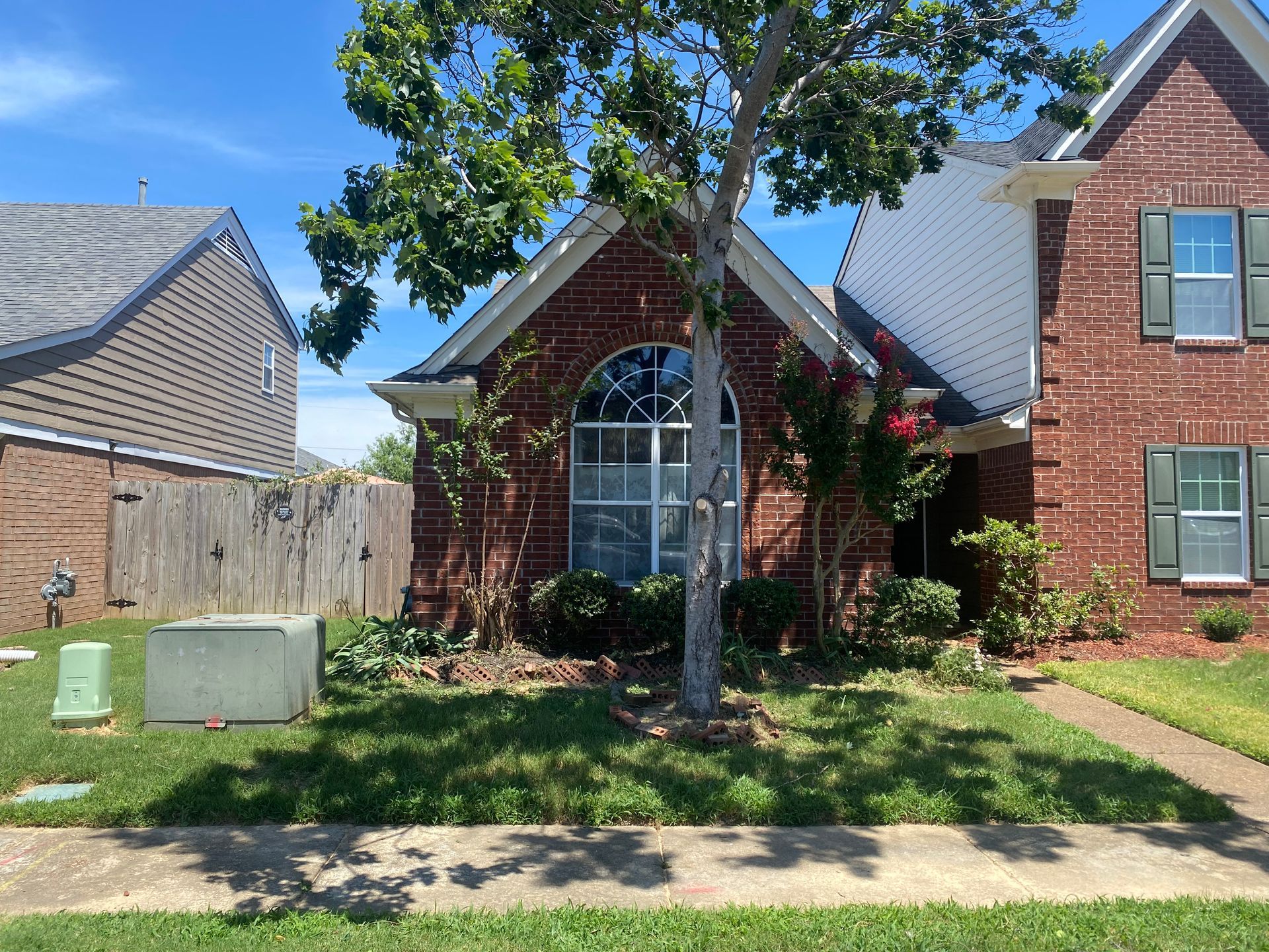 A brick house with a tree in front, green grass, and blue sky. The house has a large arched window.
