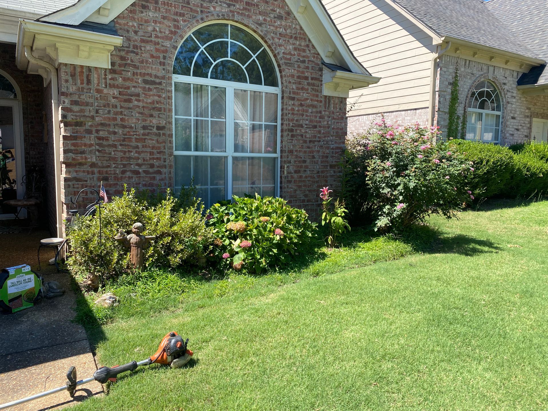 A brick house with a curved window and landscaping, a string trimmer lying on the lawn.