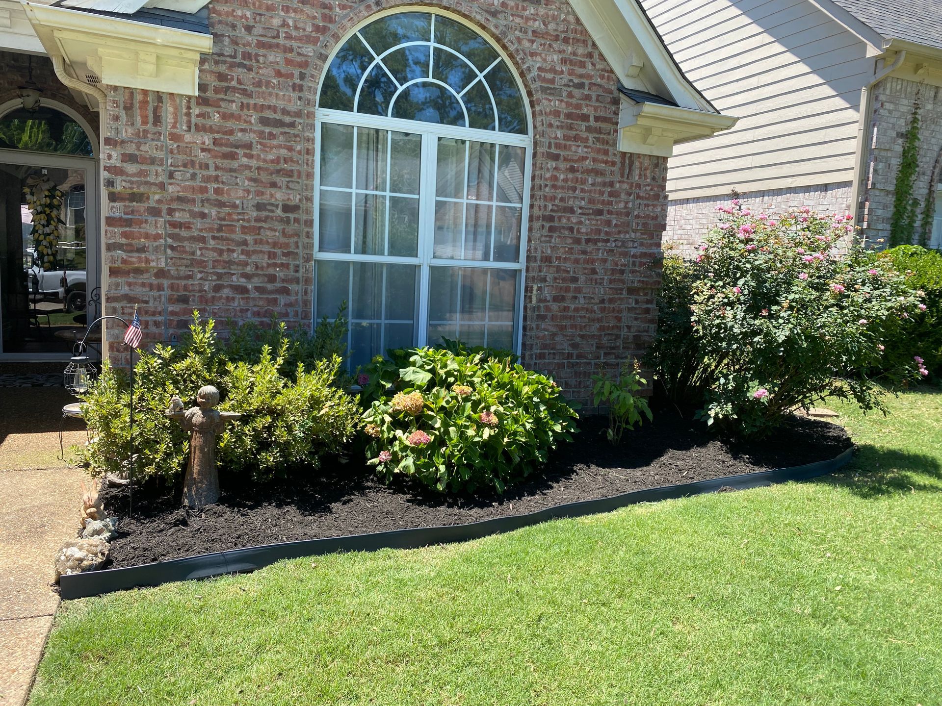 Brick house with a manicured lawn and flowerbeds, including green bushes and a window with white trim.