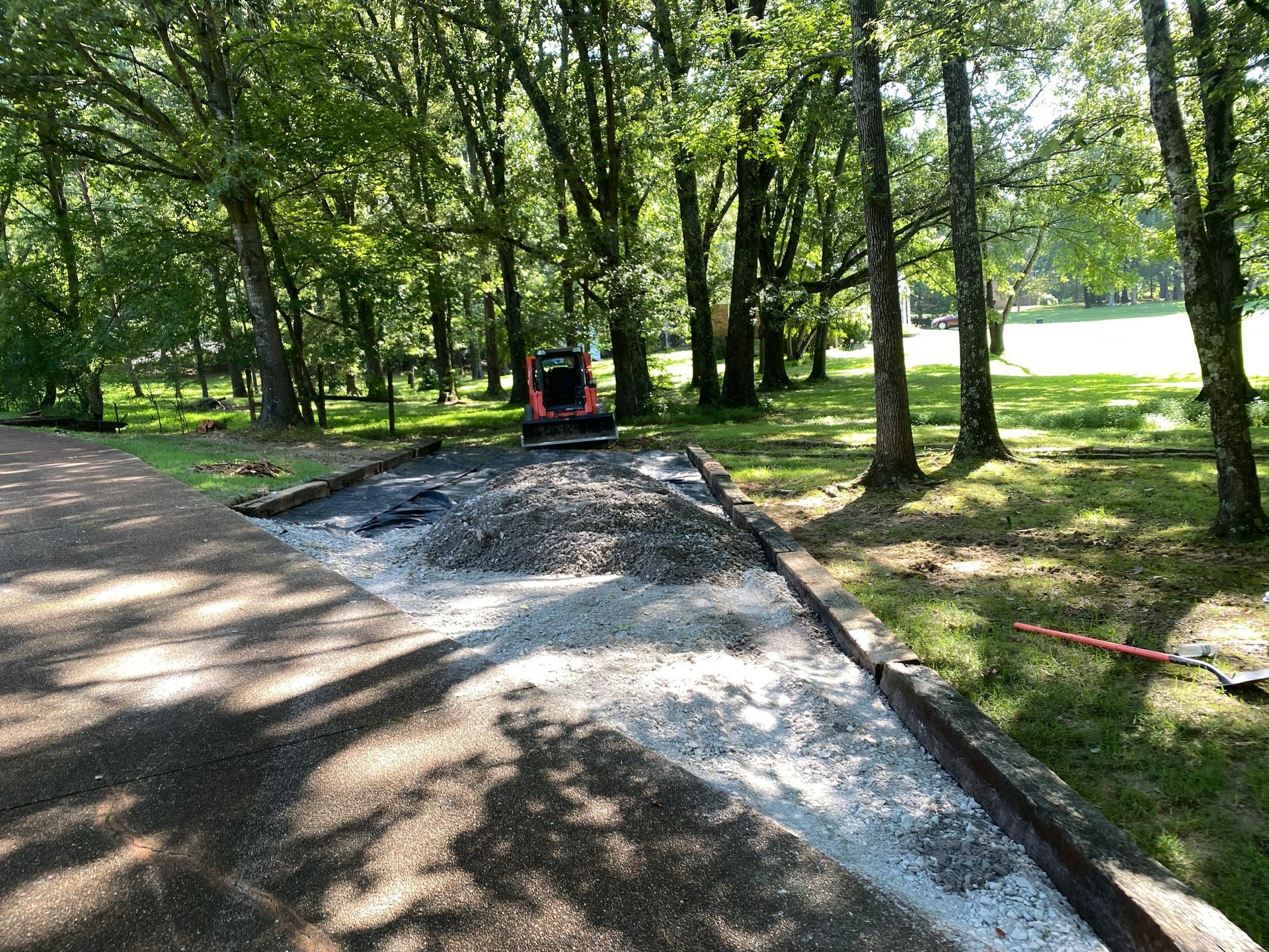 A red skid-steer loader on gravel in a tree-lined area. Construction on a path with a dark border is in progress.