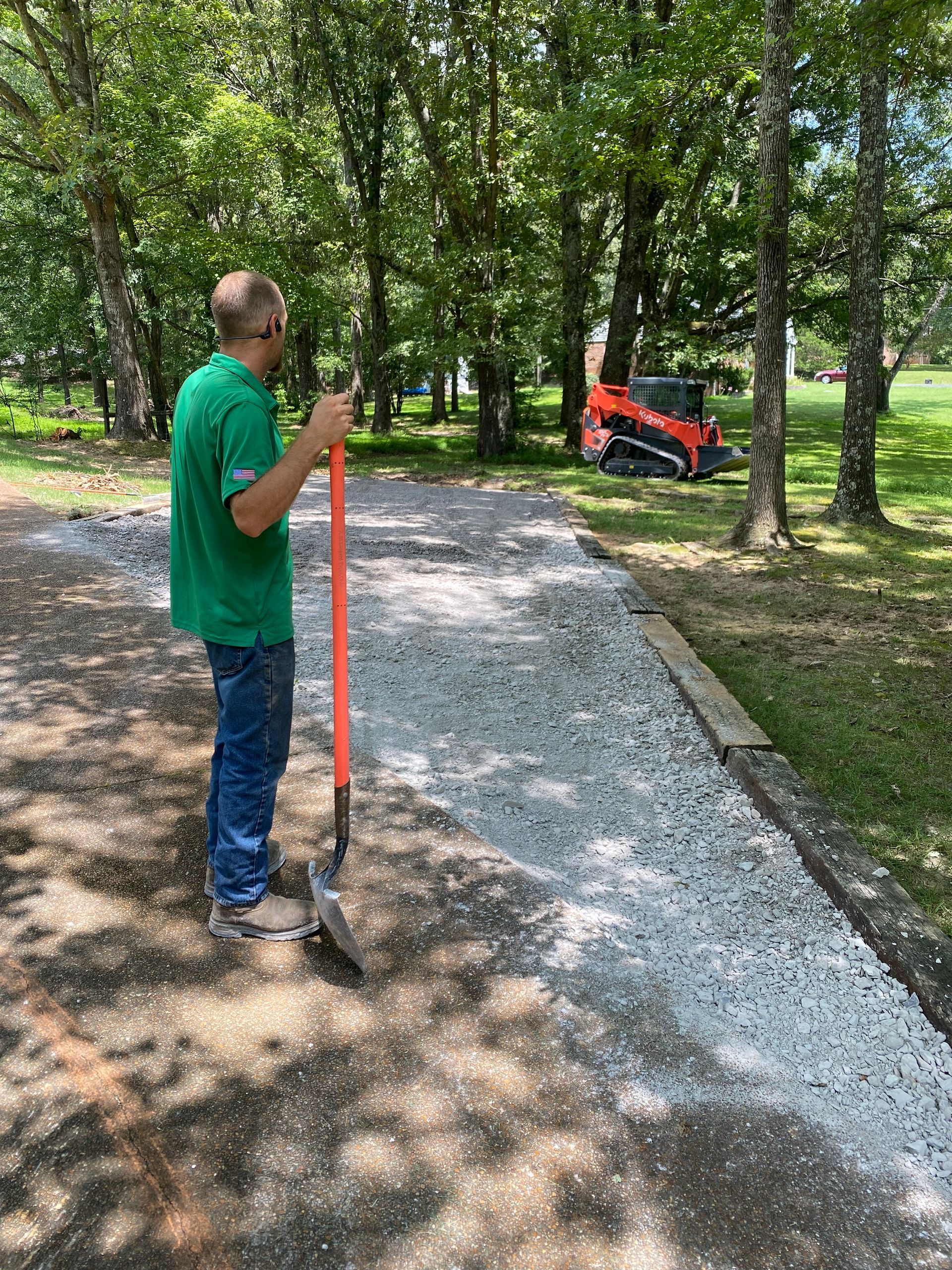 Man in green shirt raking gravel on a path. A small orange tractor and trees are in the background.