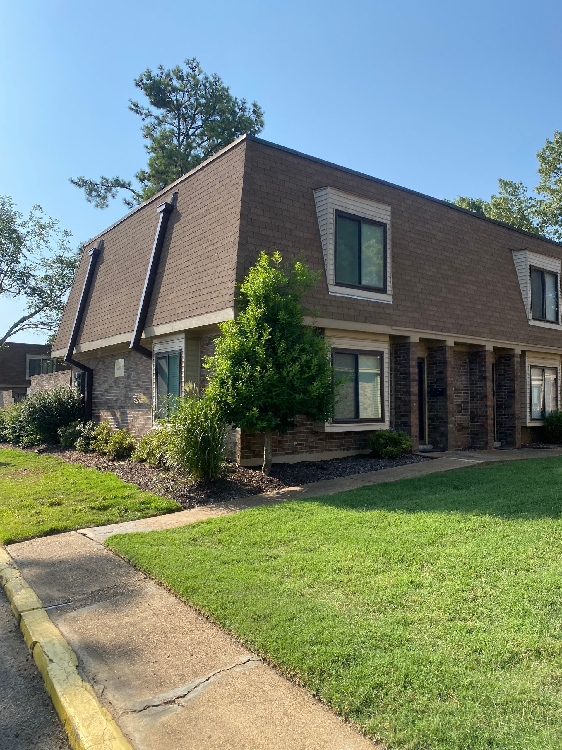 Two-story brick townhome with brown roof and green lawn. A tree is in front of the building, and trees are visible in the background.