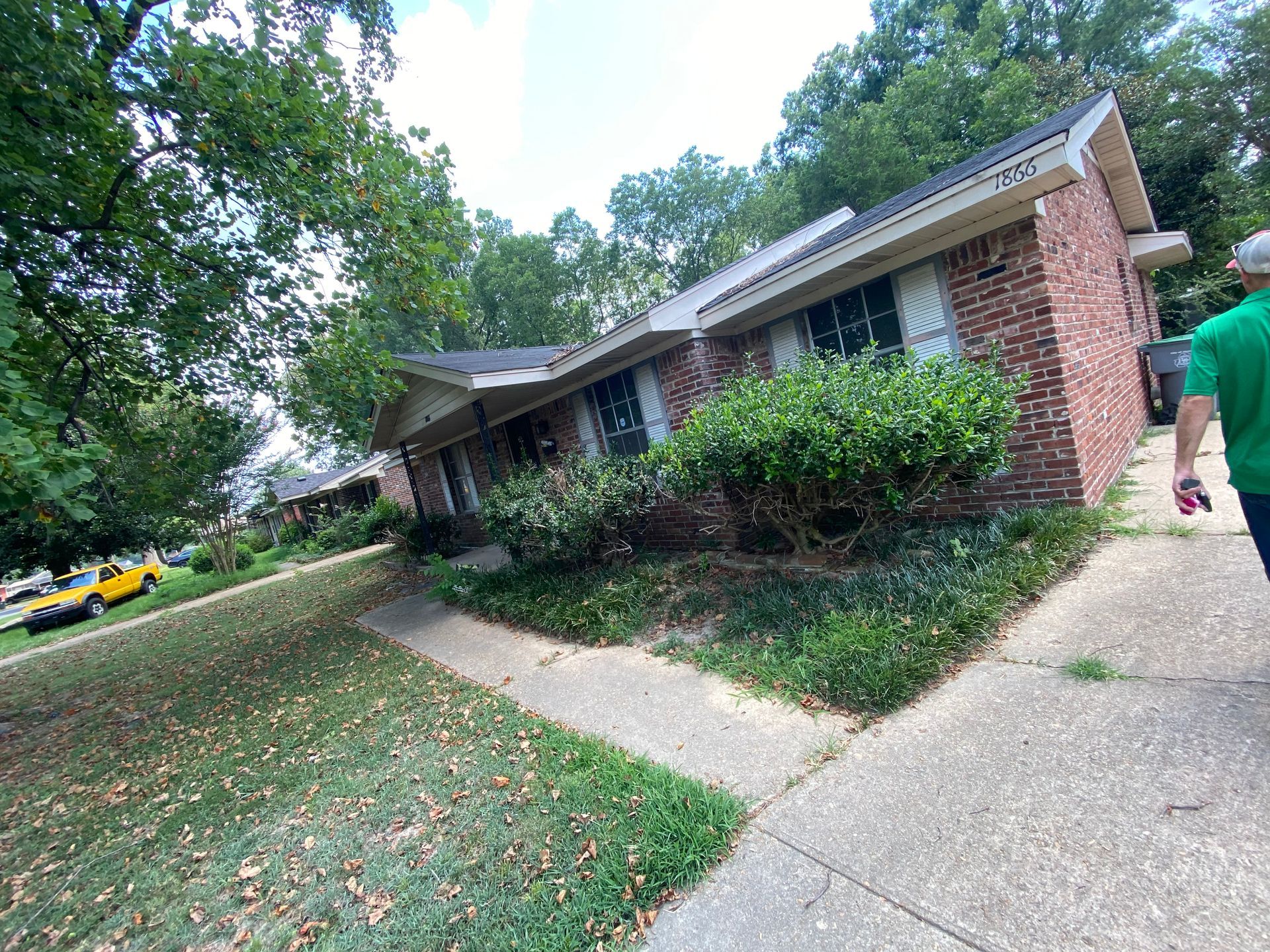 Brick house with sloped roof, surrounded by bushes and trees, sidewalk in front; person in green shirt walks away.